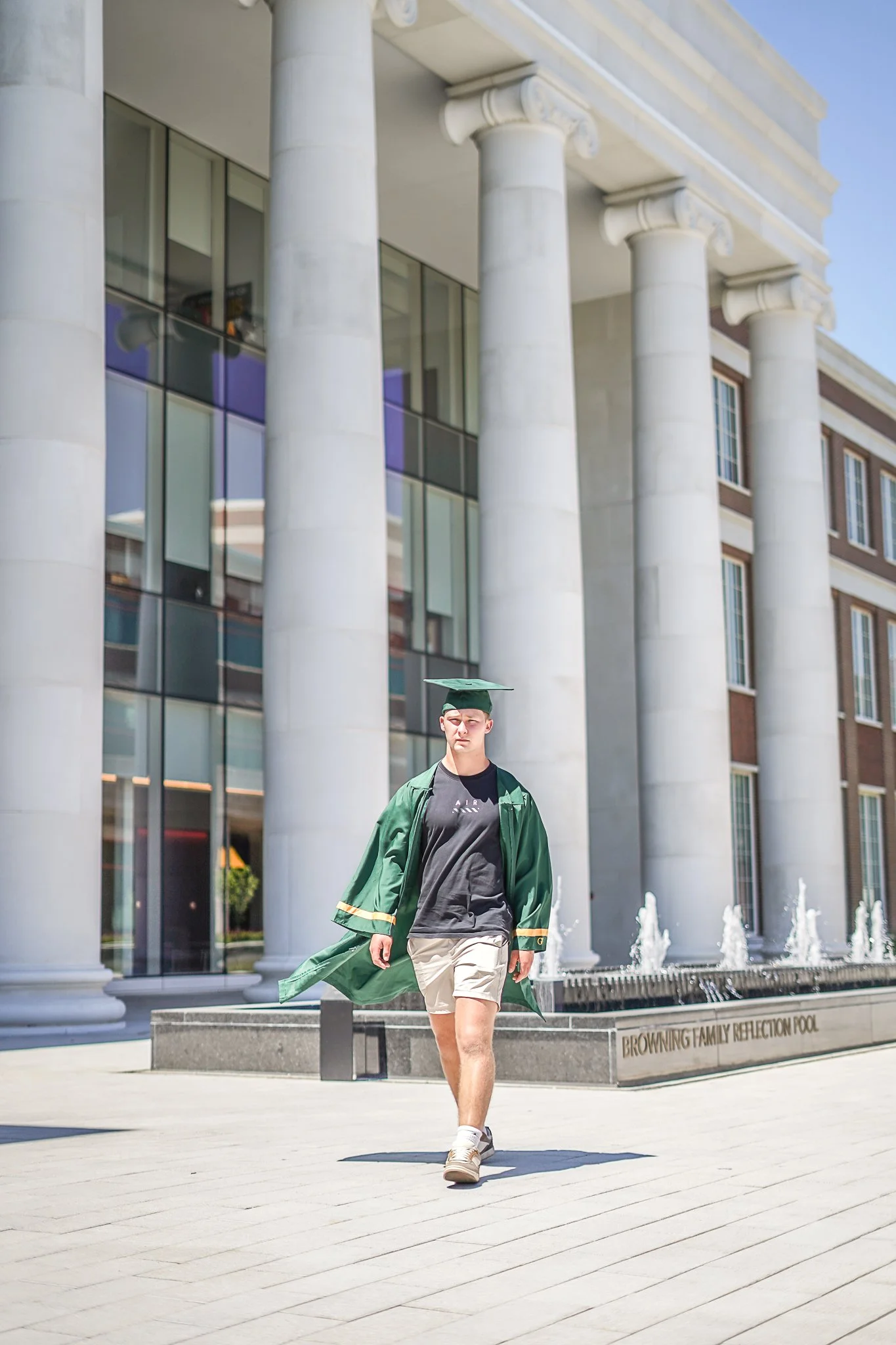 A young man in a green graduation gown and cap walking outdoors in front of a building with tall white columns and glass windows.