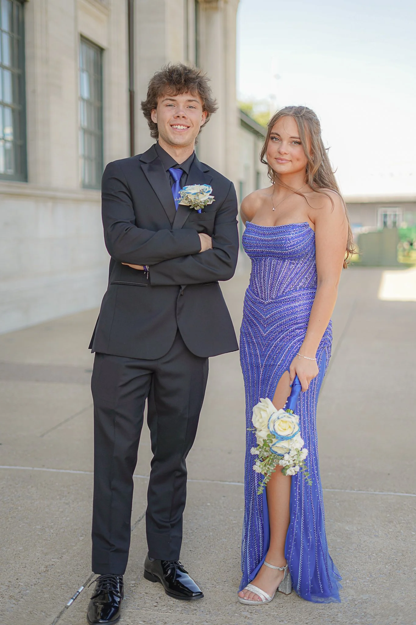 A young man in a black suit with a blue tie and a woman in a long, strapless, blue dress with silver embellishments, both standing outdoors in front of a building, holding a bouquet of white and blue roses, smiling at the camera.