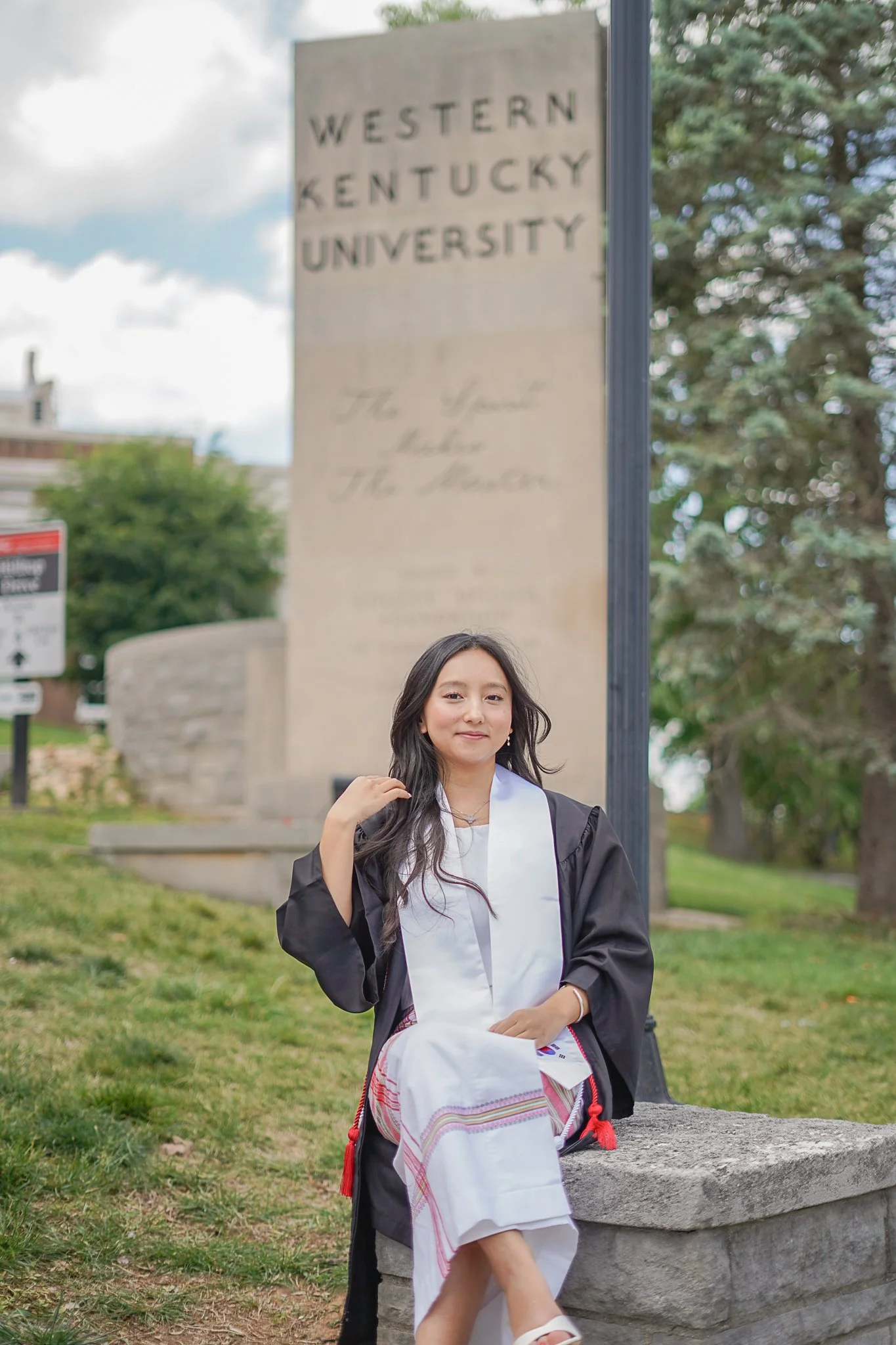 A young woman in a graduation gown and white dress sitting outdoors on a stone bench, with a large sign in the background that reads Western Kentucky University, partially obscured by a black post. She has long dark hair and is smiling at the camera.