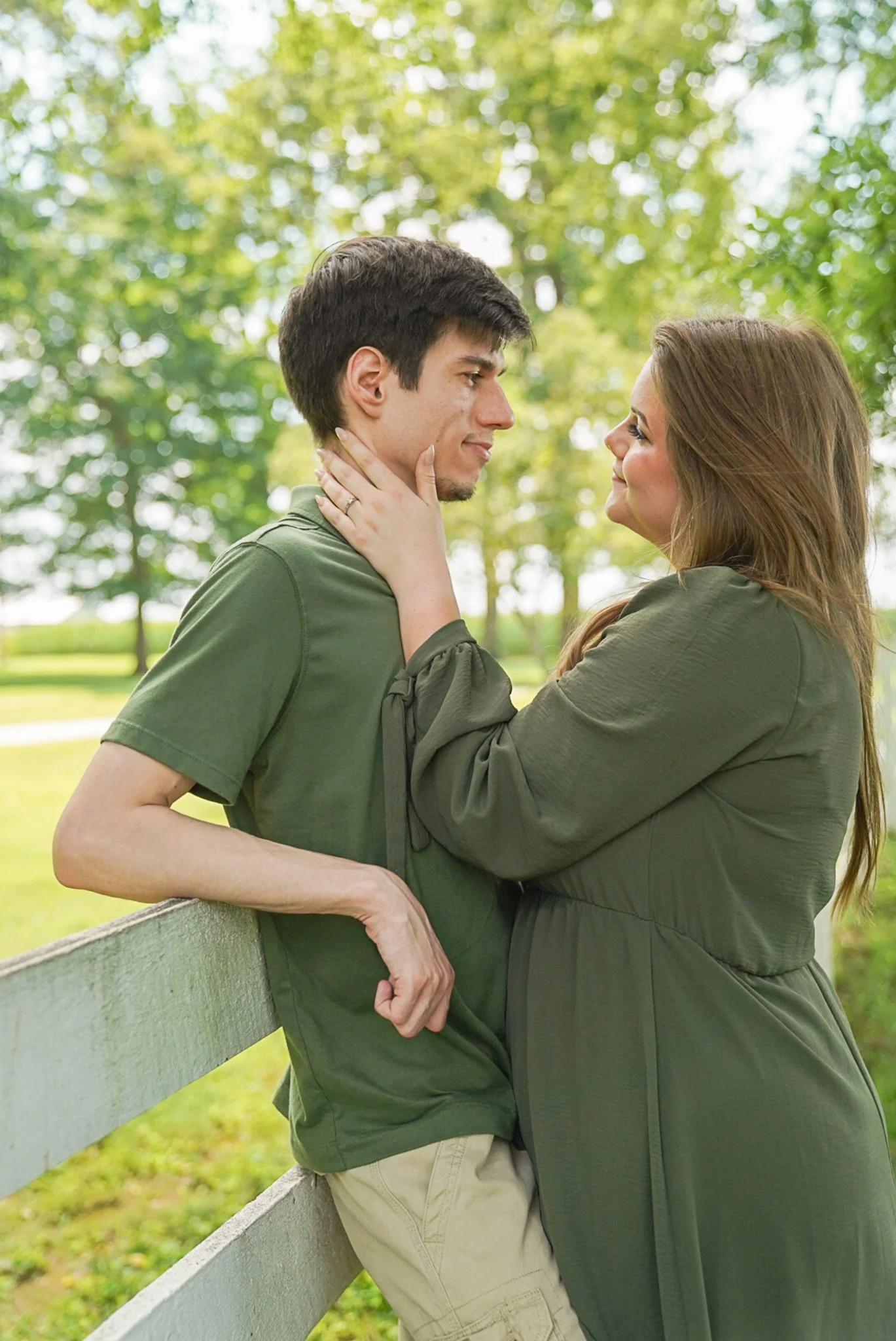 A man and a woman stand facing each other outdoors, with the woman touching the man's face gently. They are near a white wooden fence and surrounded by green trees.