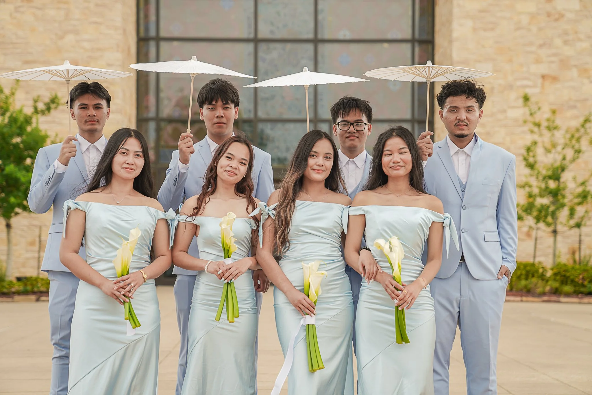 A group of nine young adults standing outside in front of a building with a large window, dressed legally in winter or summer clothes, holding white umbrellas, and flowers, smiling and posing for a photo.