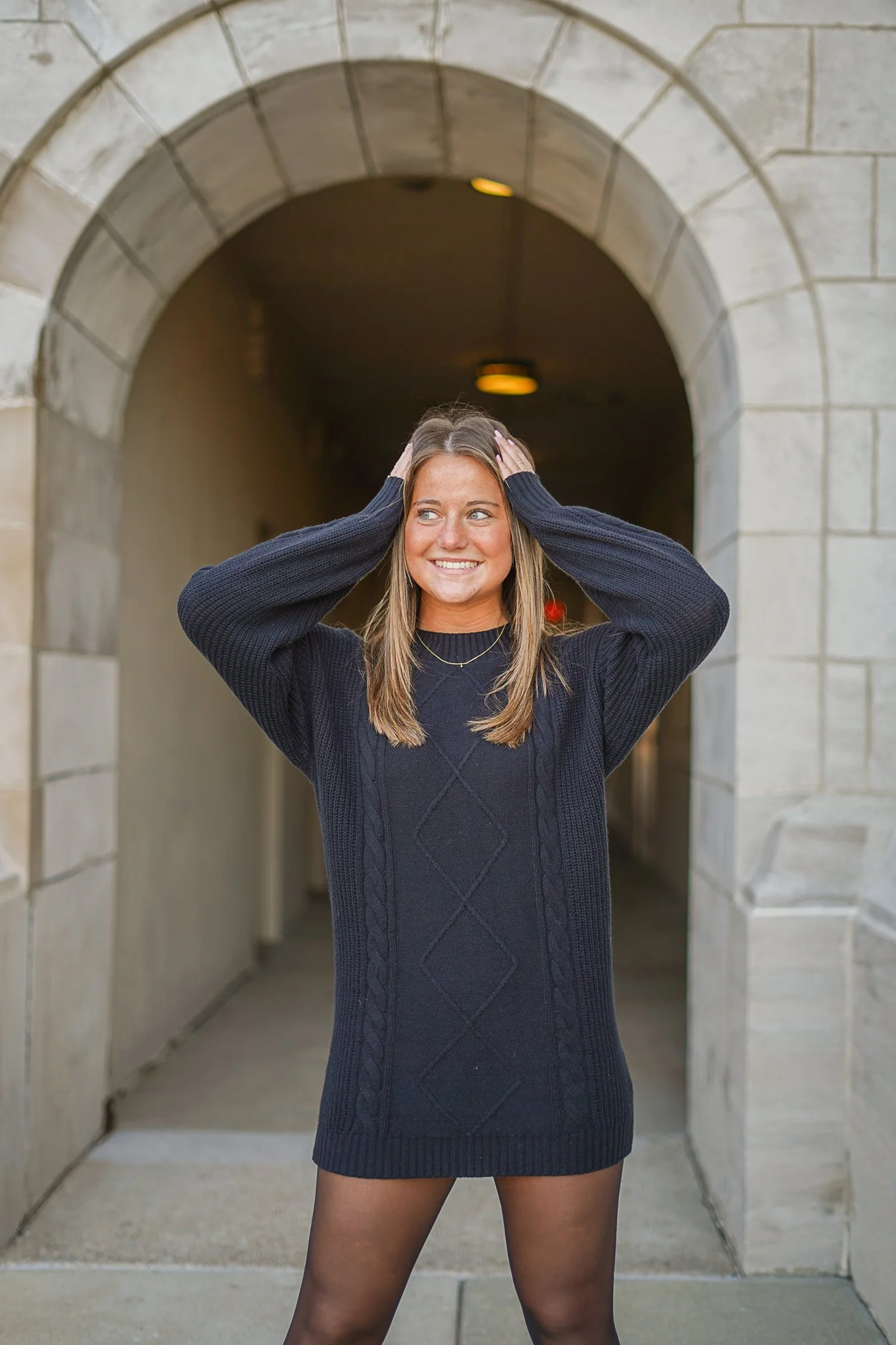 A woman with long brown hair, wearing a black sweater dress and black sheer tights, standing in front of a stone archway, smiling and holding her hands to her head.