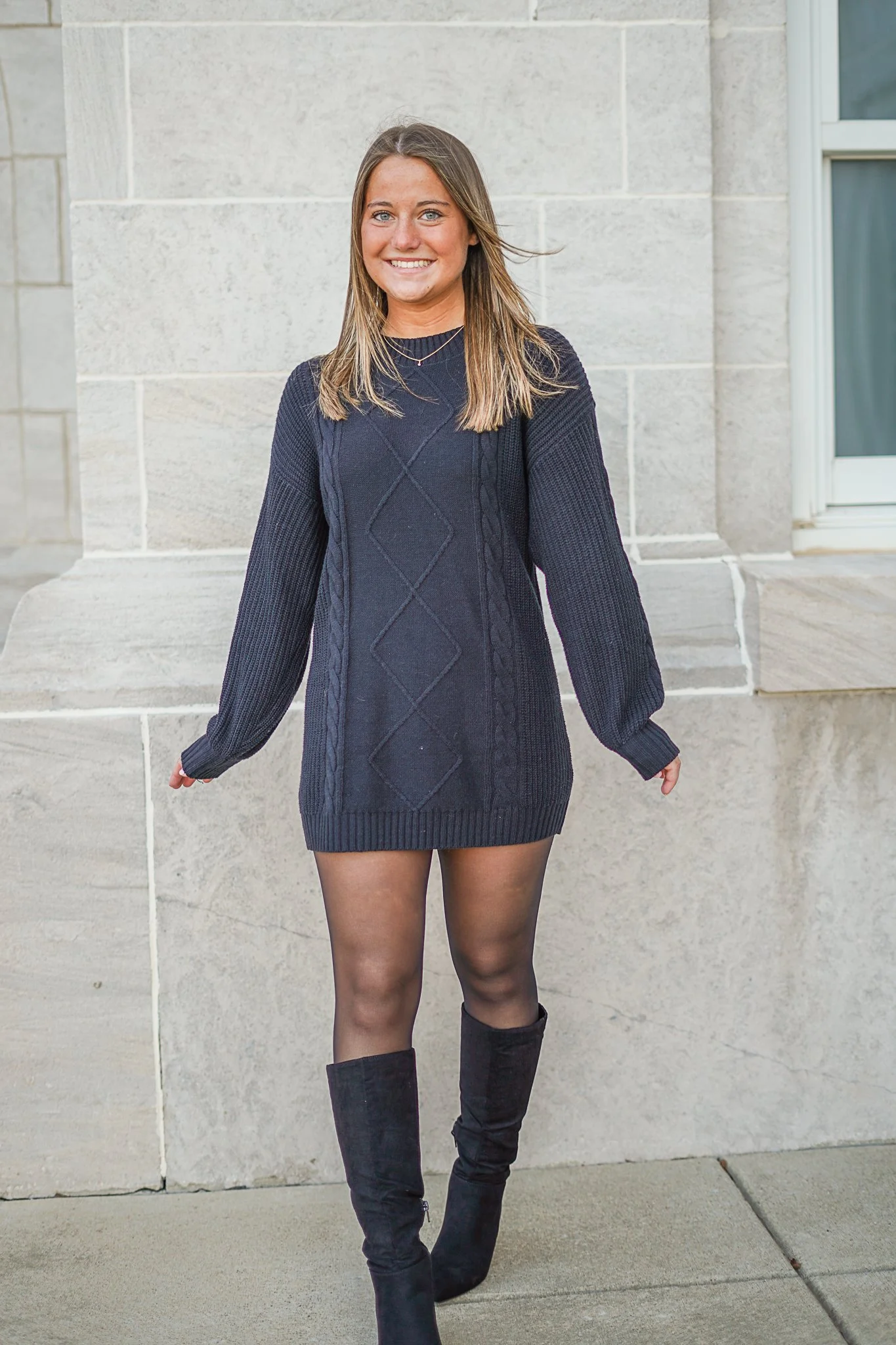 A young woman with light brown hair, wearing a navy blue sweater dress and black over-the-knee boots, standing outdoors in front of a light-colored stone wall, smiling at the camera.