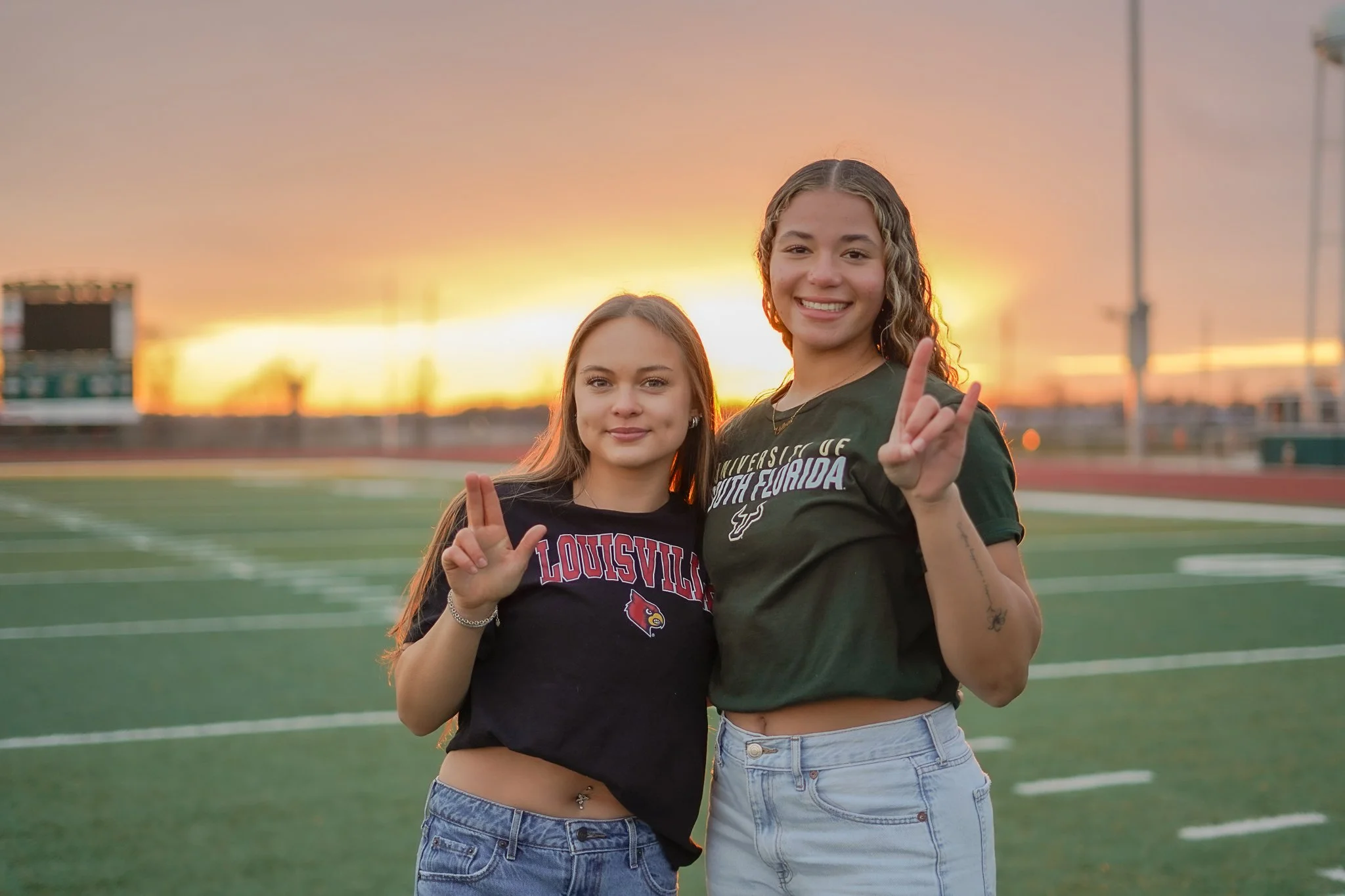 Two young women standing on a football field at sunset, smiling, making peace signs with their hands. They are wearing casual clothes and appear to be friends.