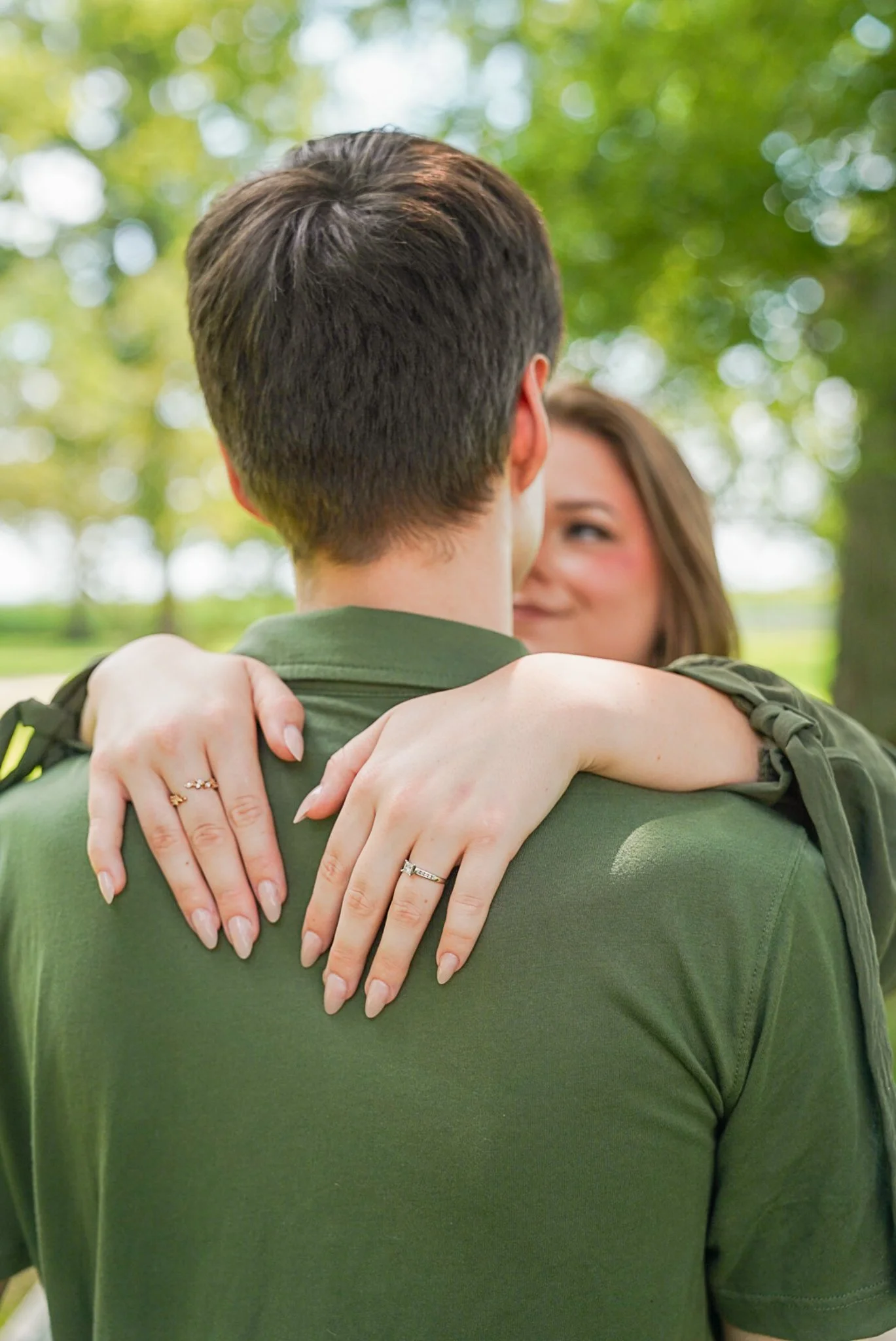 A person in a military uniform being hugged by a woman with her arms around the person's neck. The woman's face is visible, and they are outdoors with greenery in the background.