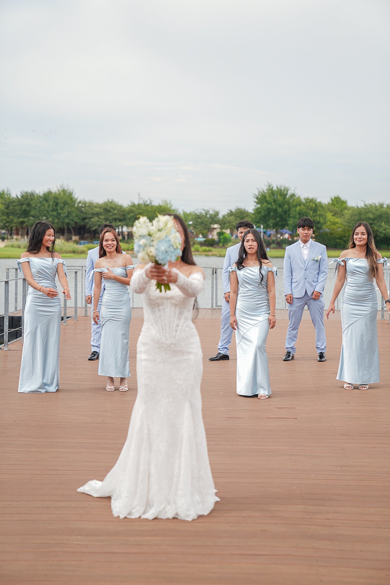 A bride in a white lace wedding gown holding a bouquet of white and blue flowers with a blurred face, standing on a wooden deck near a body of water. Several bridesmaids and groomsmen in light blue formal attire stand behind her, smiling and waiting 