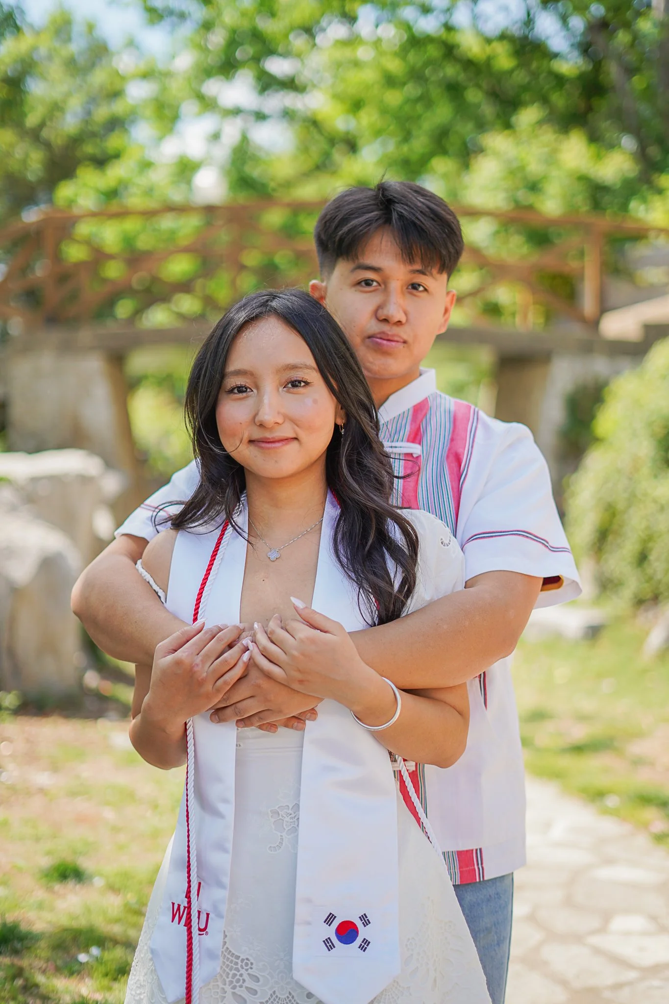A young woman and man standing outdoors in a sunny, green park. The woman is in the foreground, smiling gently, wearing a white dress with lace details and a silver necklace. The man is behind her, with his arms around her, wearing a white shirt with