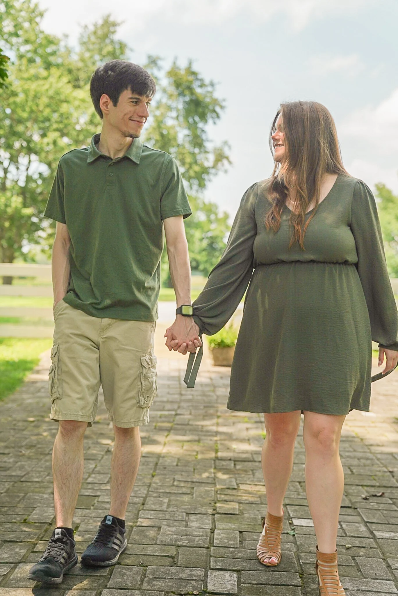 A young man and woman are walking hand in hand outdoors on a paved path with trees and a cloudy sky in the background. The man is wearing a green polo shirt, beige cargo shorts, and black sneakers. The woman is wearing a dark green dress, tan high-he