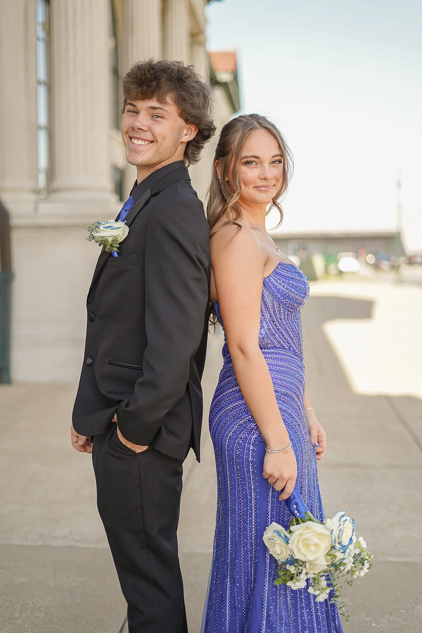 Young couple dressed in formal attire, standing back-to-back outdoors, with the man in a black suit and the woman in a blue gown, holding a bouquet of white roses with blue accents.