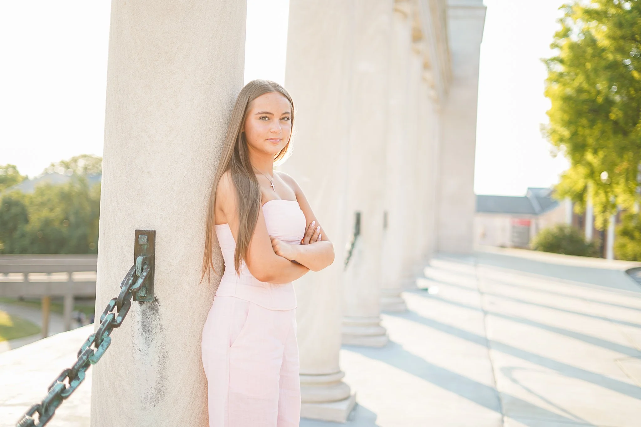 Young woman with long hair and a necklace standing outside, leaning against a large white column with chains attached, with a building and green trees in the background.