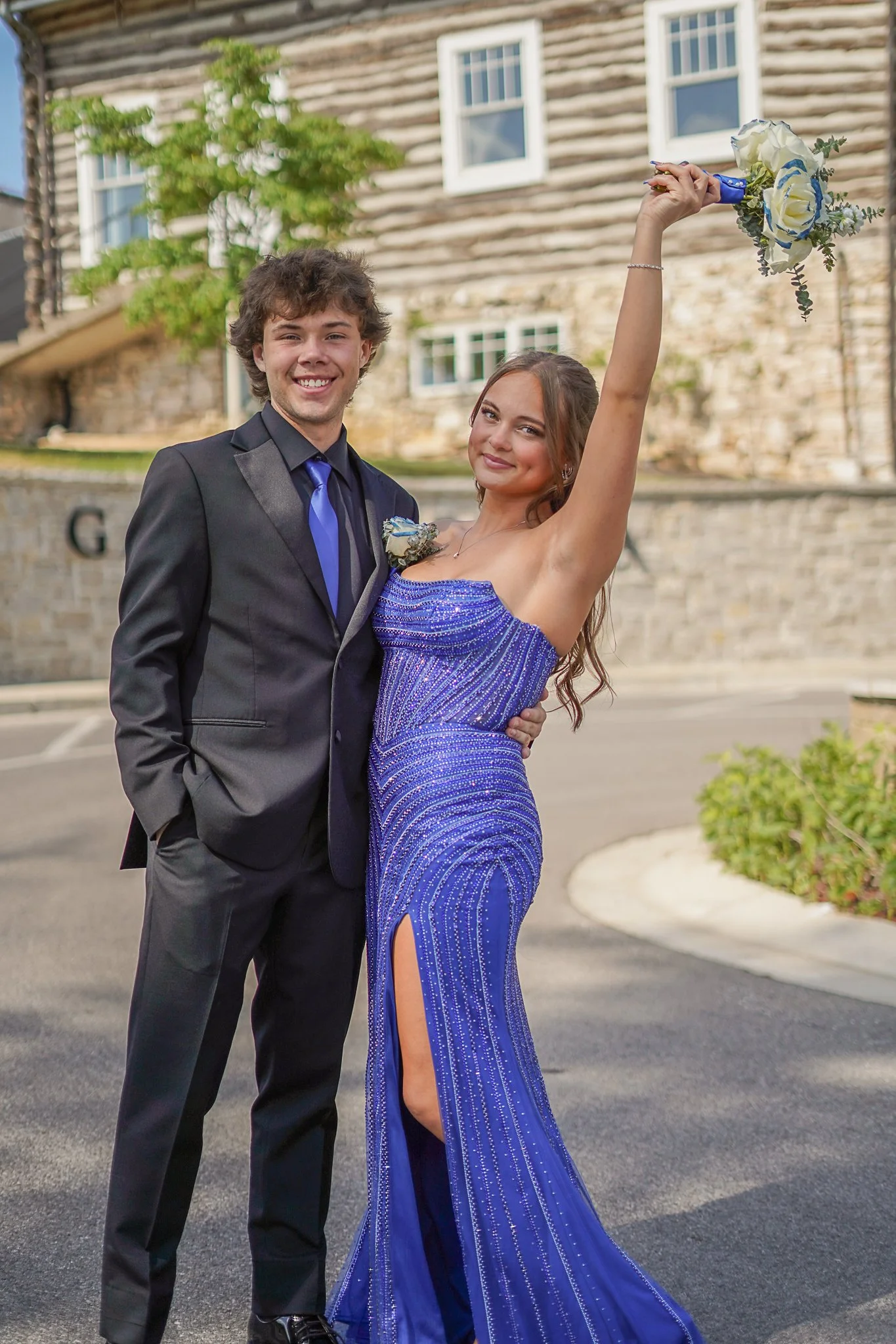 A young man in a black tuxedo with a blue tie and a young woman in a blue sequined gown holding a bouquet, standing outside in front of a building with railroad ties and windows, smiling and posing for a photo.