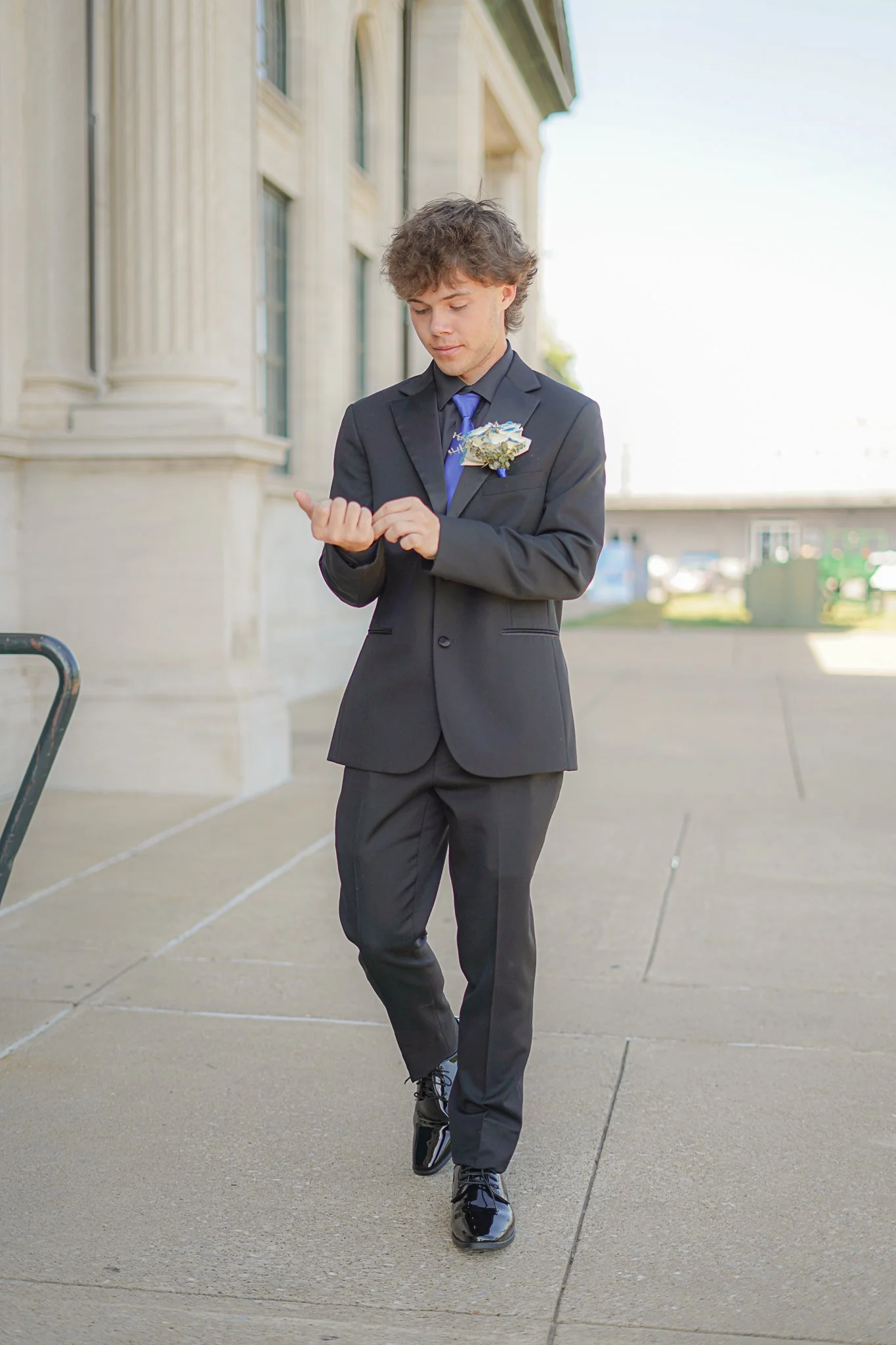 Young man in a black suit with a blue tie and boutonniere, walking on city sidewalk, looking at his watch.