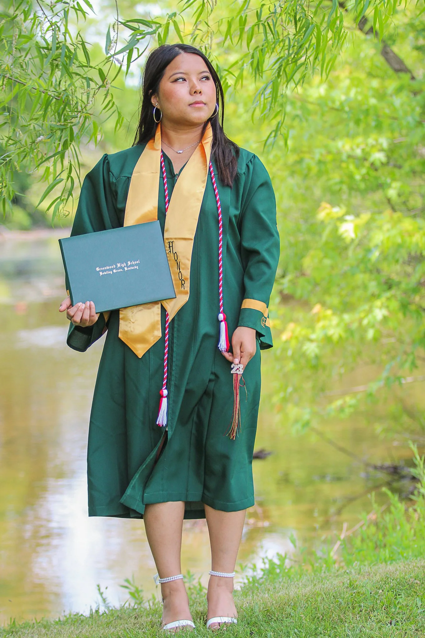 A woman in a green graduation gown and yellow stole stands outdoors near a body of water, holding a diploma, with trees and greenery in the background.