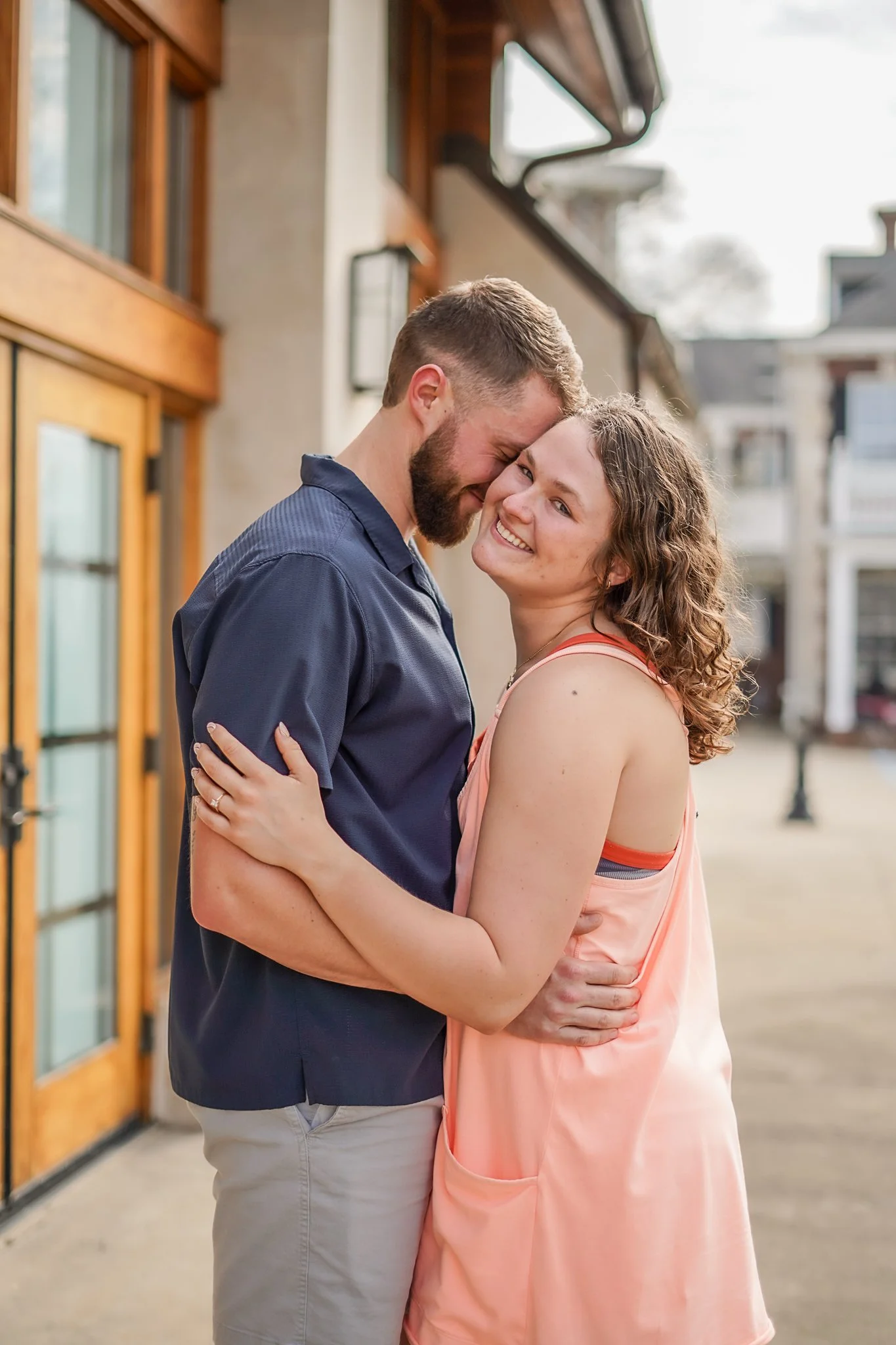 A young couple embraces intimately outside a building with wooden and brick elements, smiling affectionately at each other.