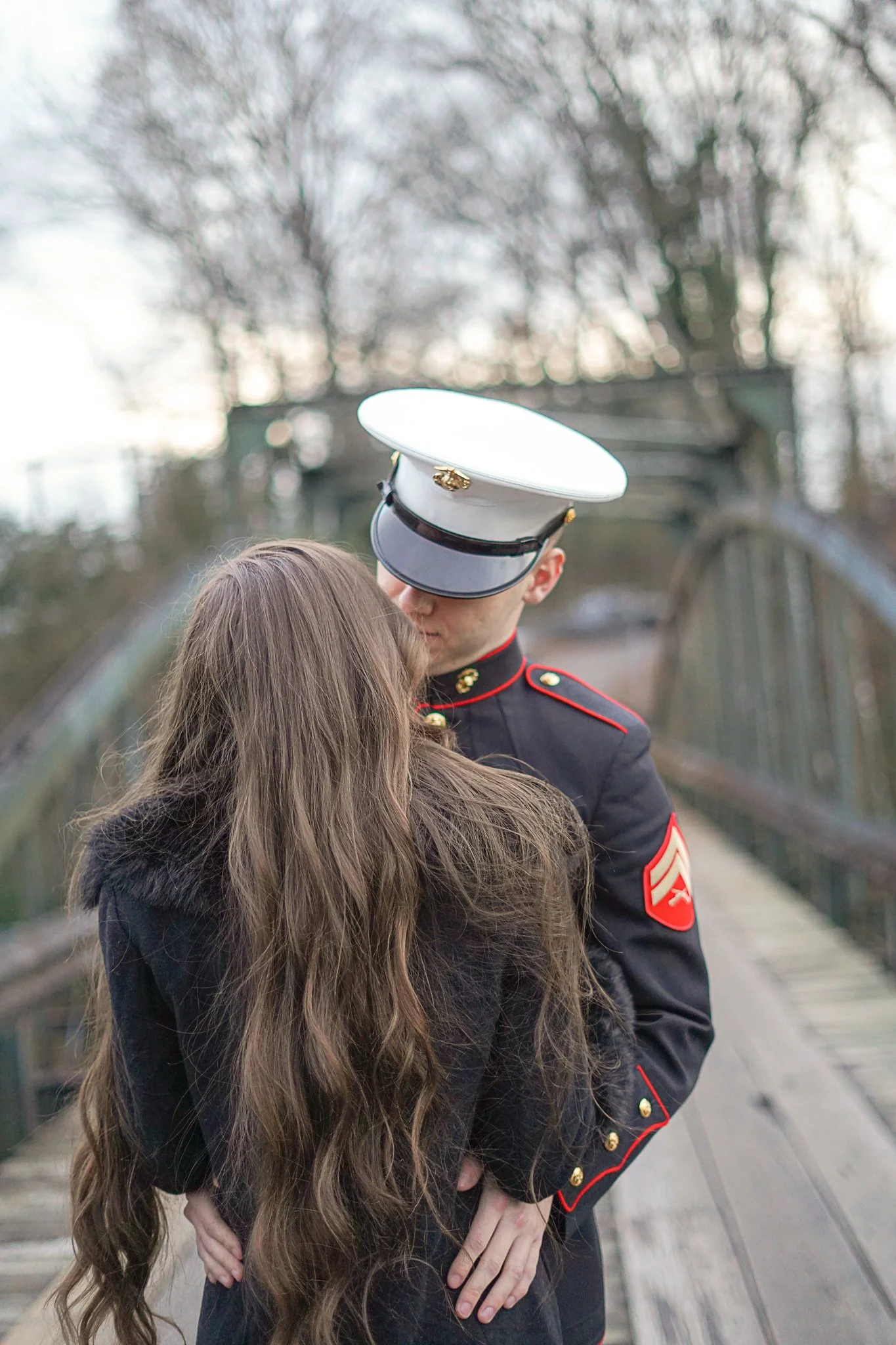 A person with long brown hair, wearing a black jacket, is standing close to a Marine dressed in a formal military uniform with a white hat, on a bridge with a blurred wooded background.