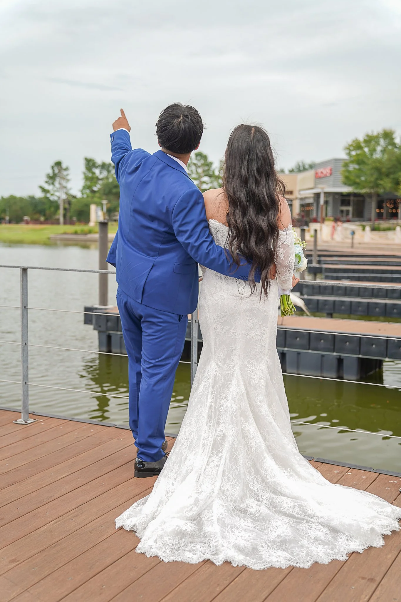 A newlywed couple standing on a wooden dock by a body of water, with the groom pointing toward the sky and the bride holding a bouquet, both dressed in wedding attire, with a small town or shopping area across the water.