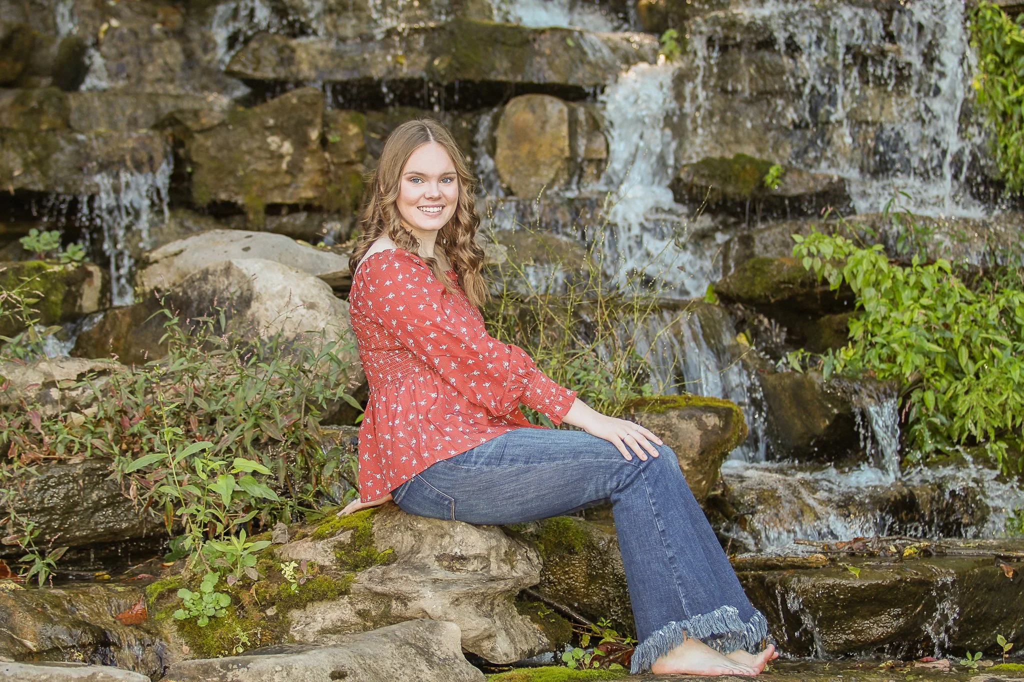 A young woman with long wavy hair smiling while sitting on rocks by a waterfall, wearing a red floral blouse and jeans, in a natural outdoor setting.