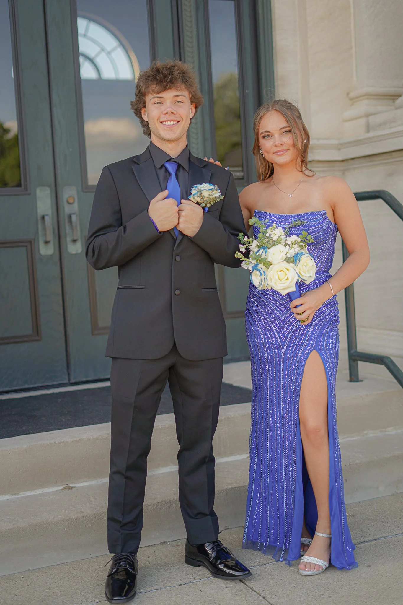 A young man and woman dressed formally, standing outside a building with glass doors. The man is in a black suit with a blue tie and boutonniere, smiling, with fists clenched near chest. The woman is in a strapless blue gown with a high slit, holding