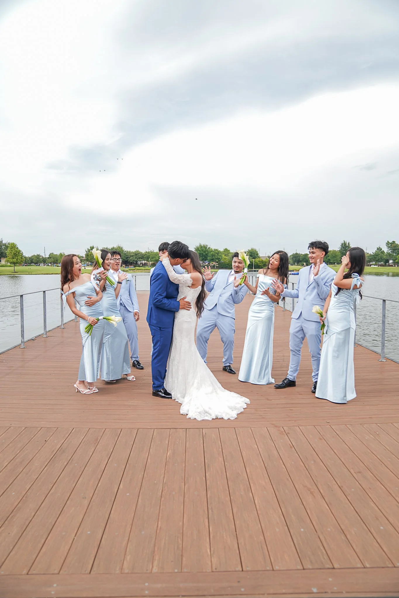 A group of people dressed in formal attire participating in a wedding ceremony on a wooden dock by the water, with a cloudy sky overhead.