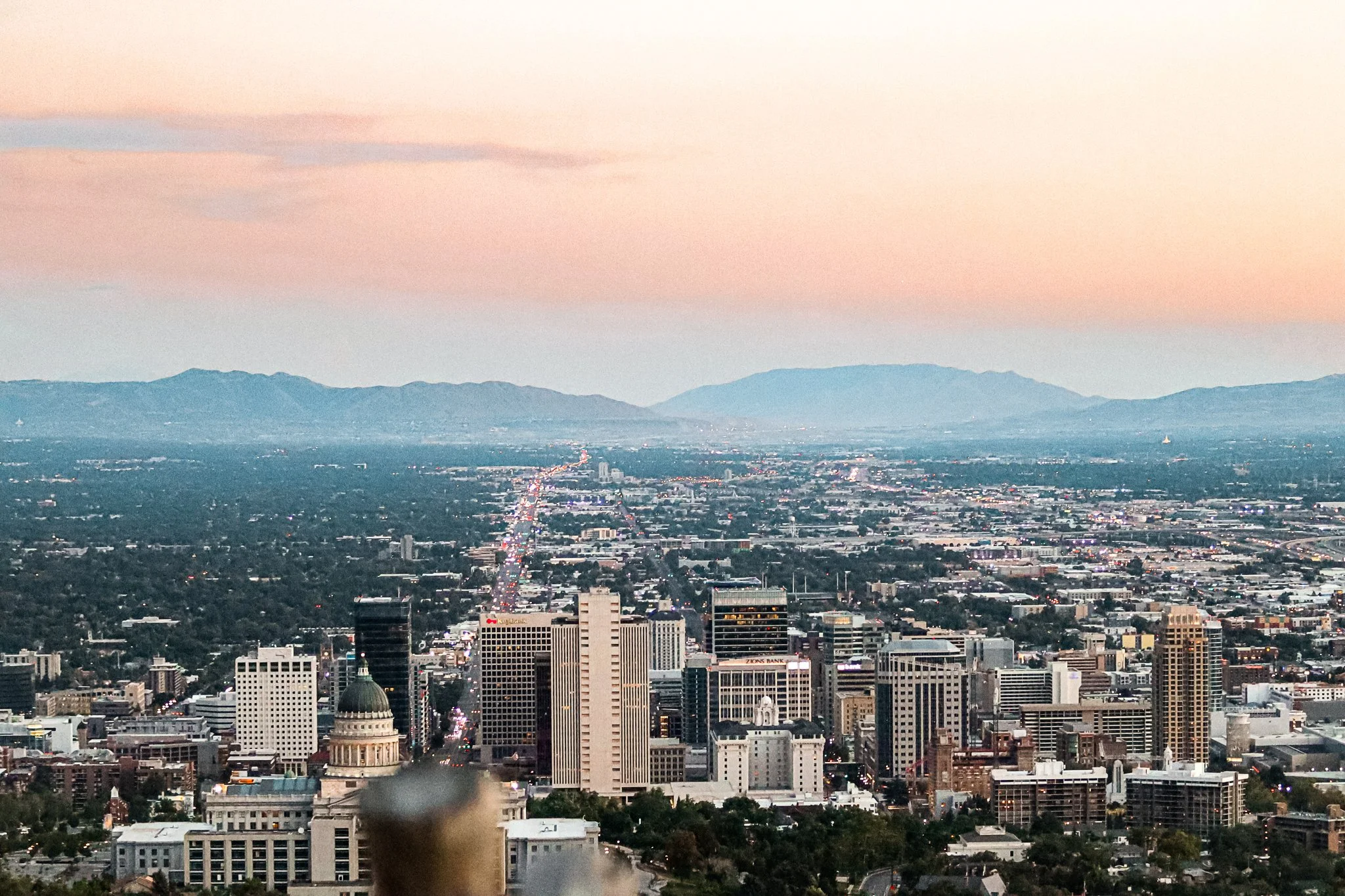 A cityscape view of downtown with skyscrapers, a distant mountain range, and a pink and blue sky during sunset or dusk.