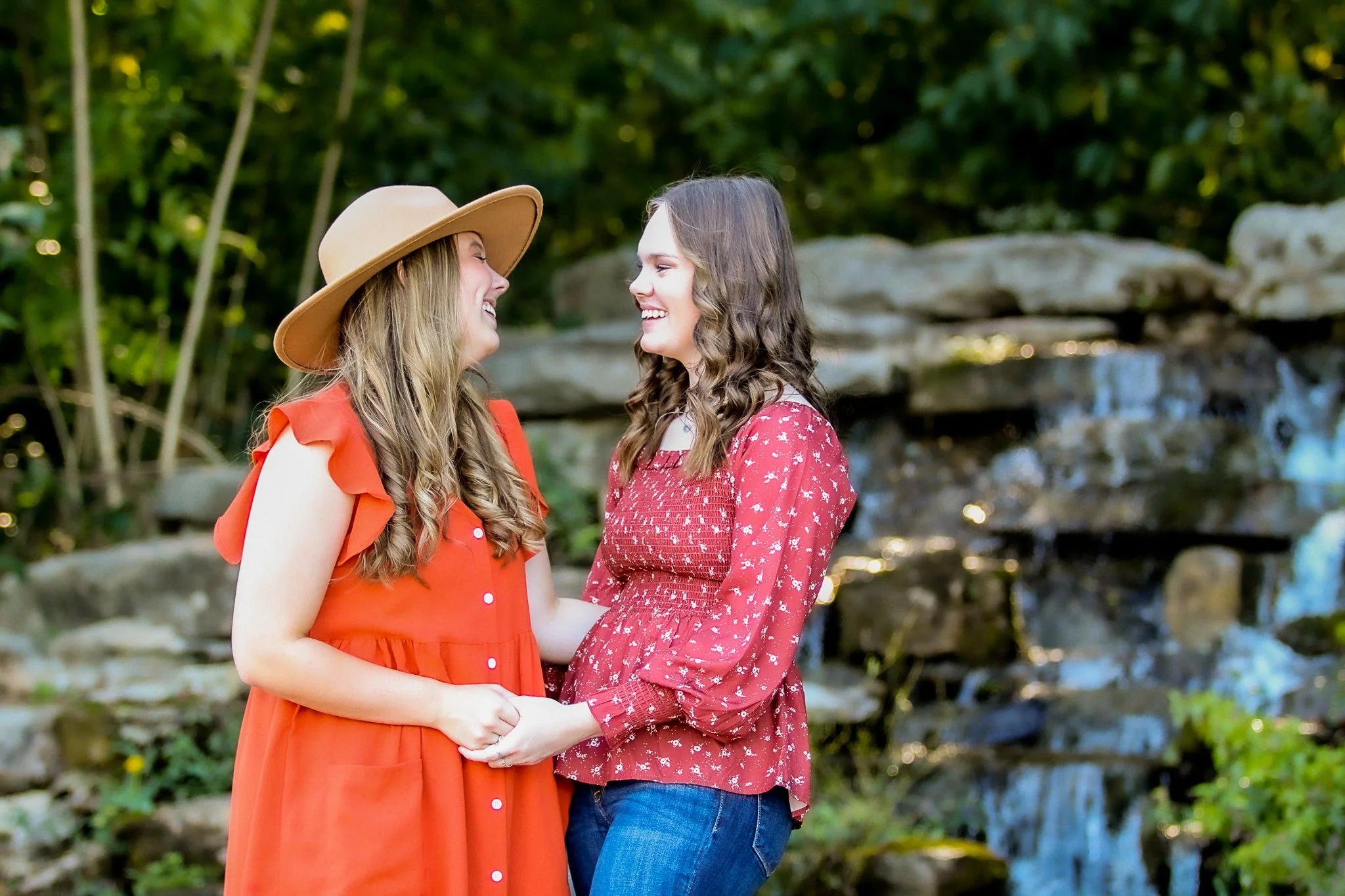 Two women smiling and holding hands outdoors near a waterfall, with lush green trees in the background.