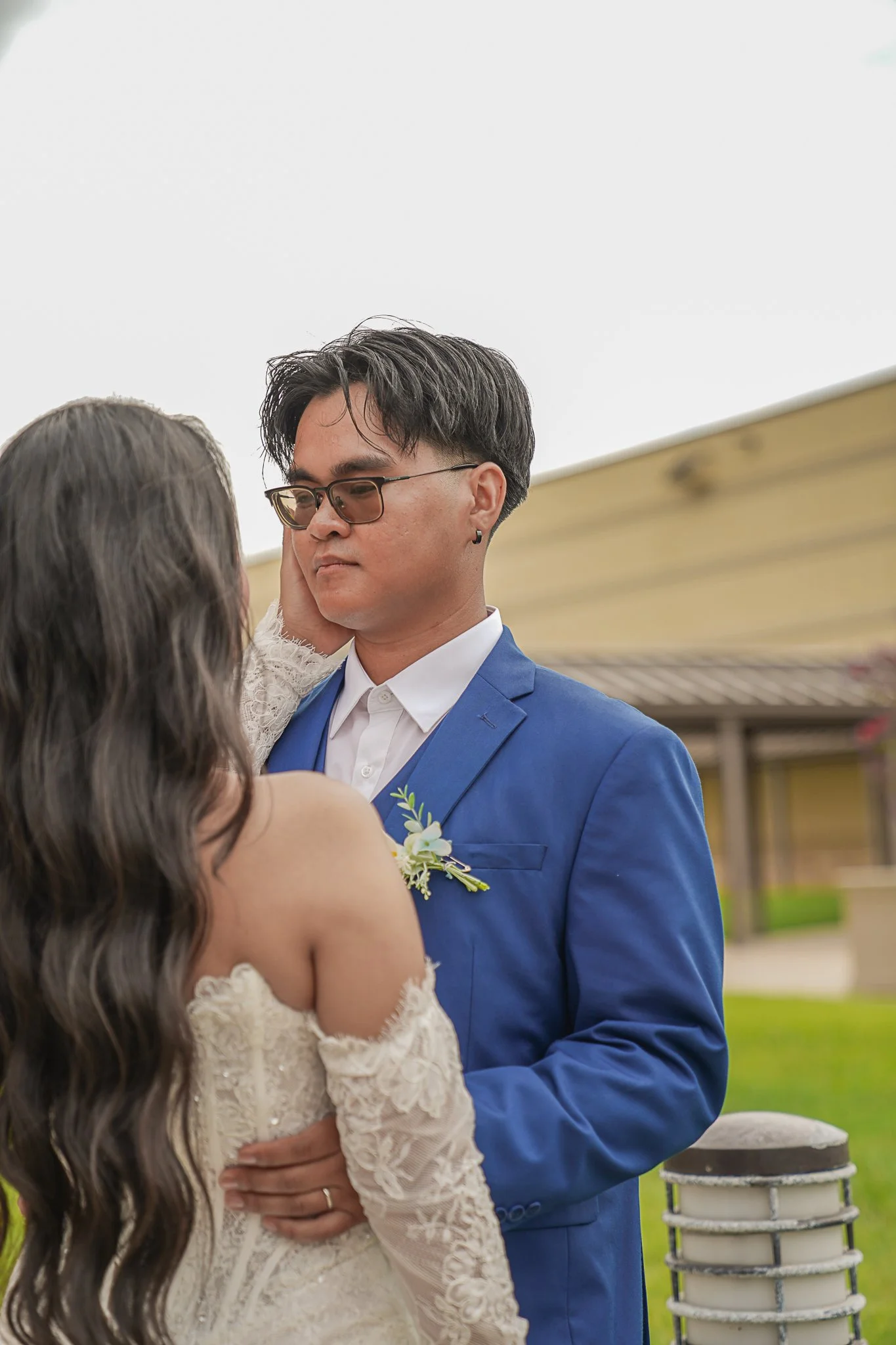 A couple sharing a kiss outdoors, with the man in a blue suit and glasses, and the woman in a white lace dress with long wavy hair.