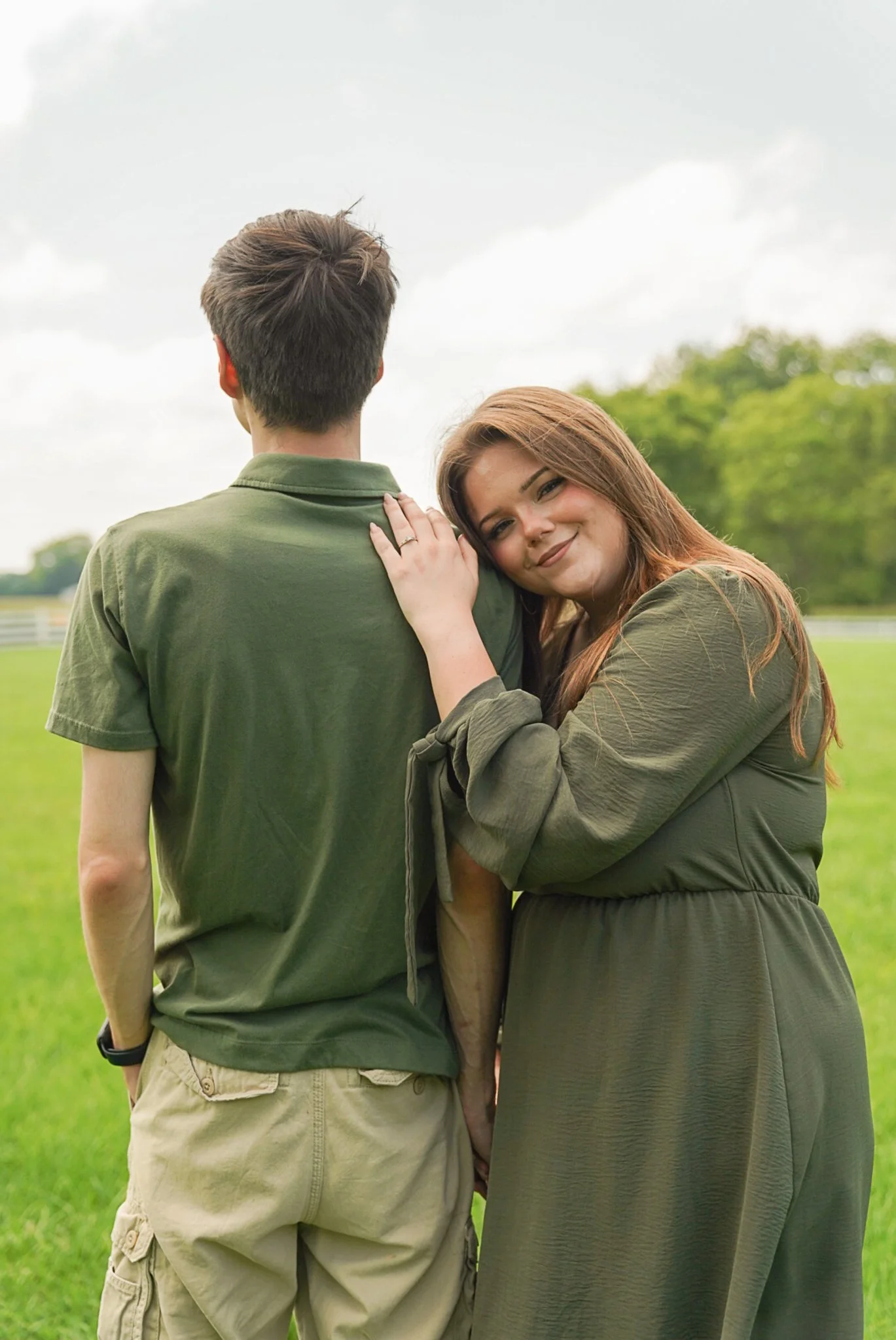 A woman with long red hair and a man with his back turned, in an outdoor grassy area with trees and a cloudy sky, woman resting her head on man's shoulder, smiling.