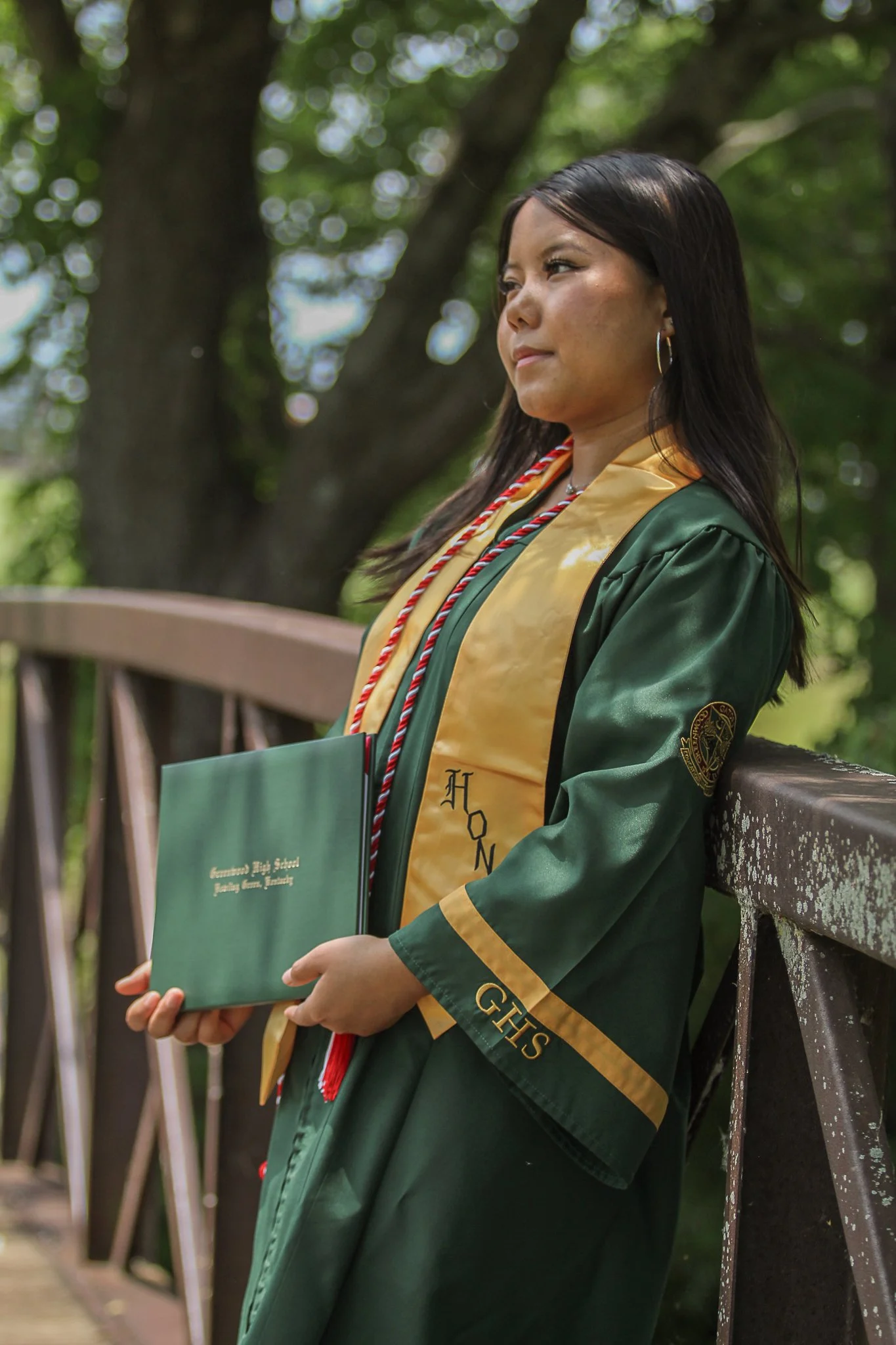 A woman in a green graduation gown and cap, holding a diploma, sitting on a park bench with trees in the background.