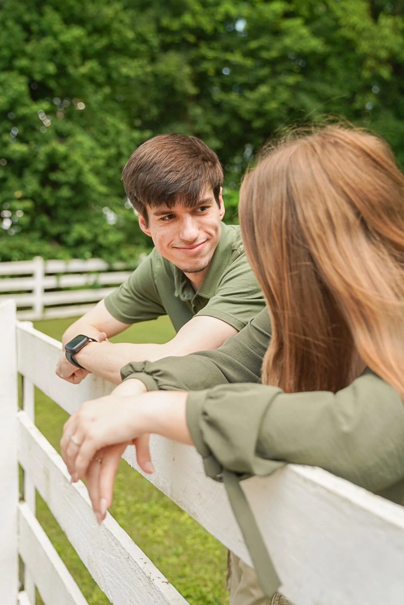 A young man and woman leaning on a white fence outdoors, engaging in conversation, with green trees in the background.