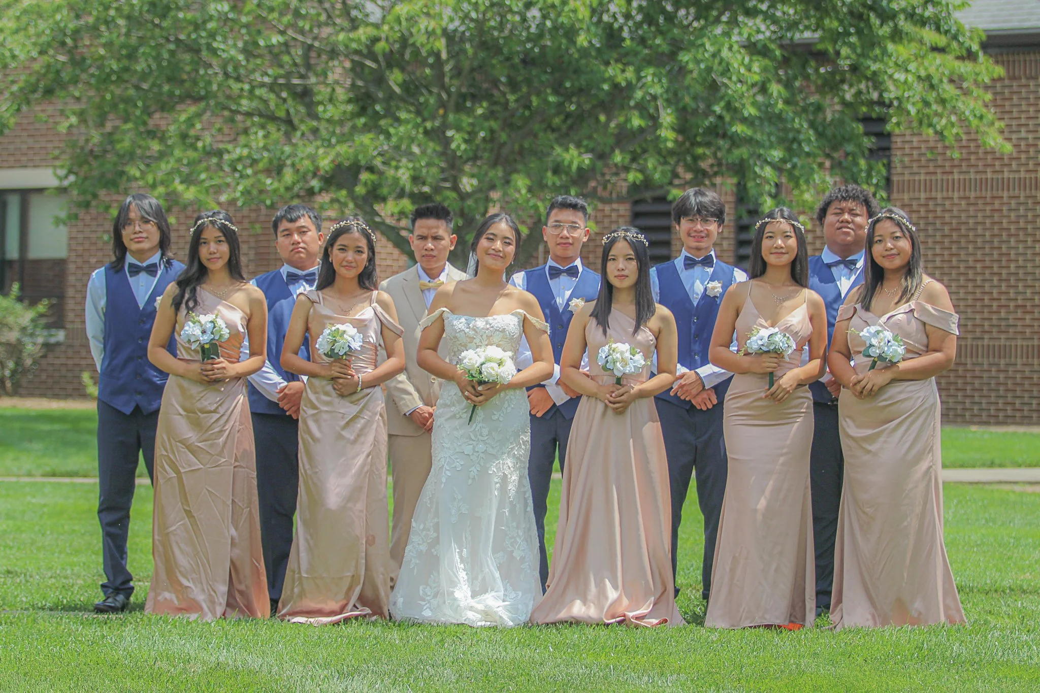 A group of twelve young people dressed in formal wedding attire standing together outdoors on grass with trees and a brick building in the background.