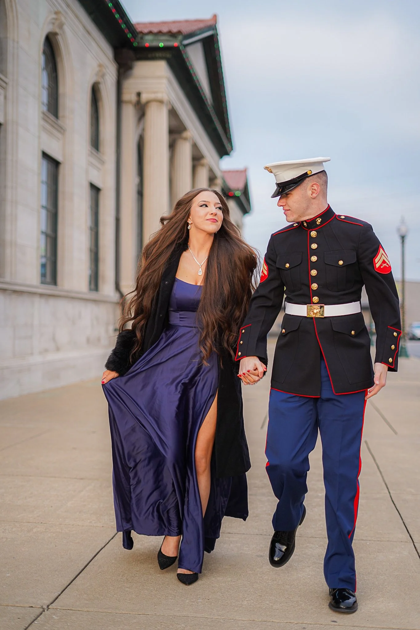Woman in a purple dress and a man in a United States Marine Corps uniform holding hands and walking together outside near a building with columns.