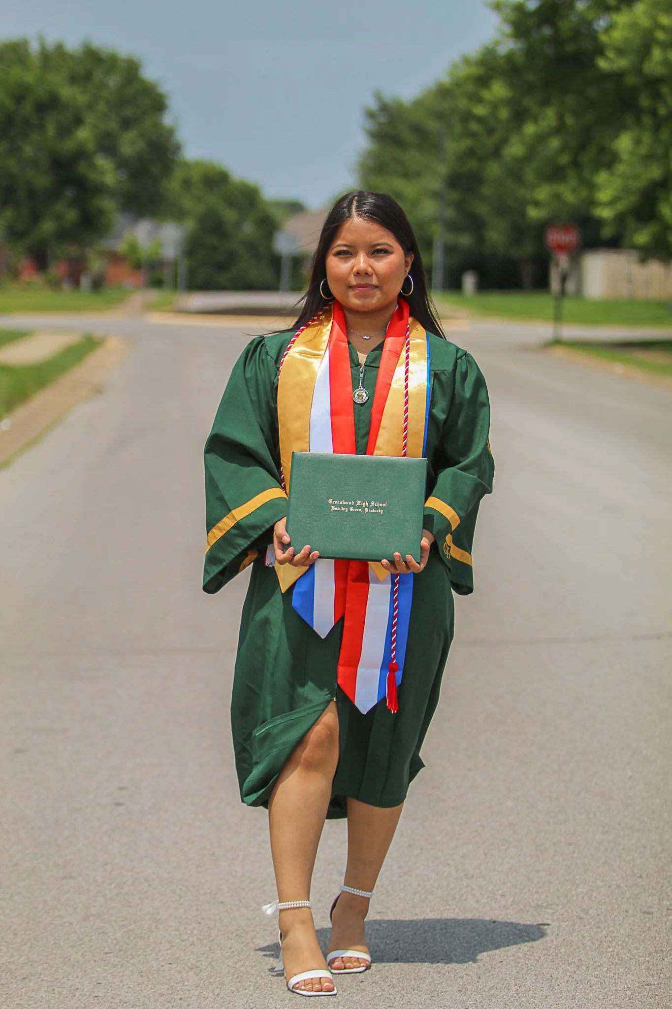 A young woman in a green graduation gown and cap holding a diploma on a suburban street, wearing white heels and standing confidently.