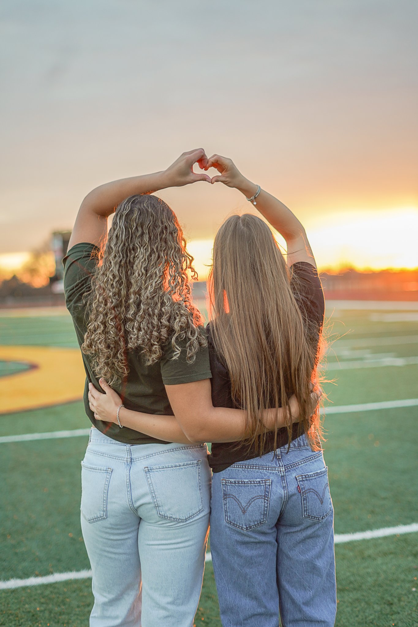 Two women hugging at a sports field during sunset, forming a heart shape with their hands.