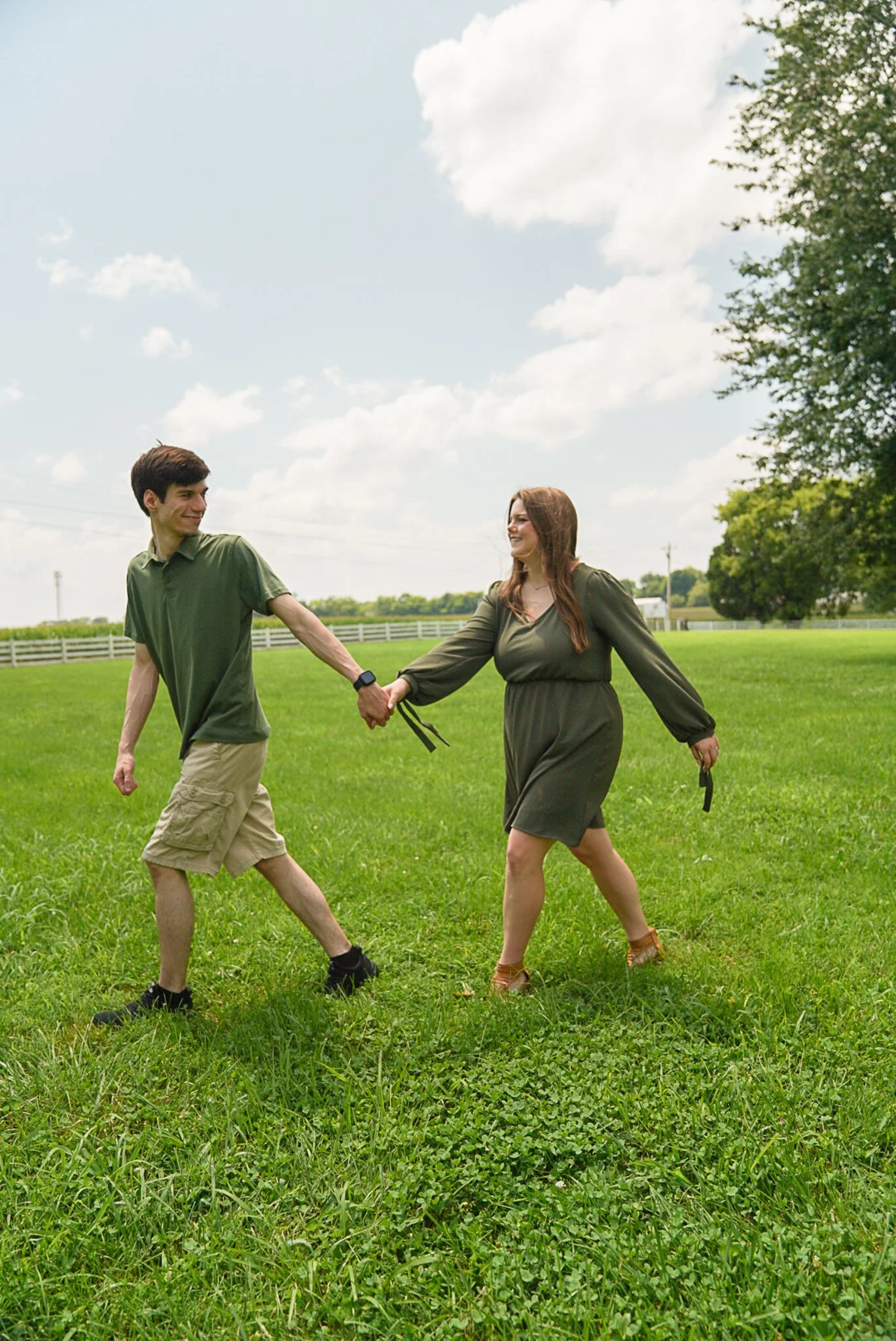 A man and a woman holding hands and walking in a green field on a sunny day. The man is wearing a green shirt and beige shorts, and the woman is wearing a dark green dress and brown sandals. Both are smiling and looking at each other.