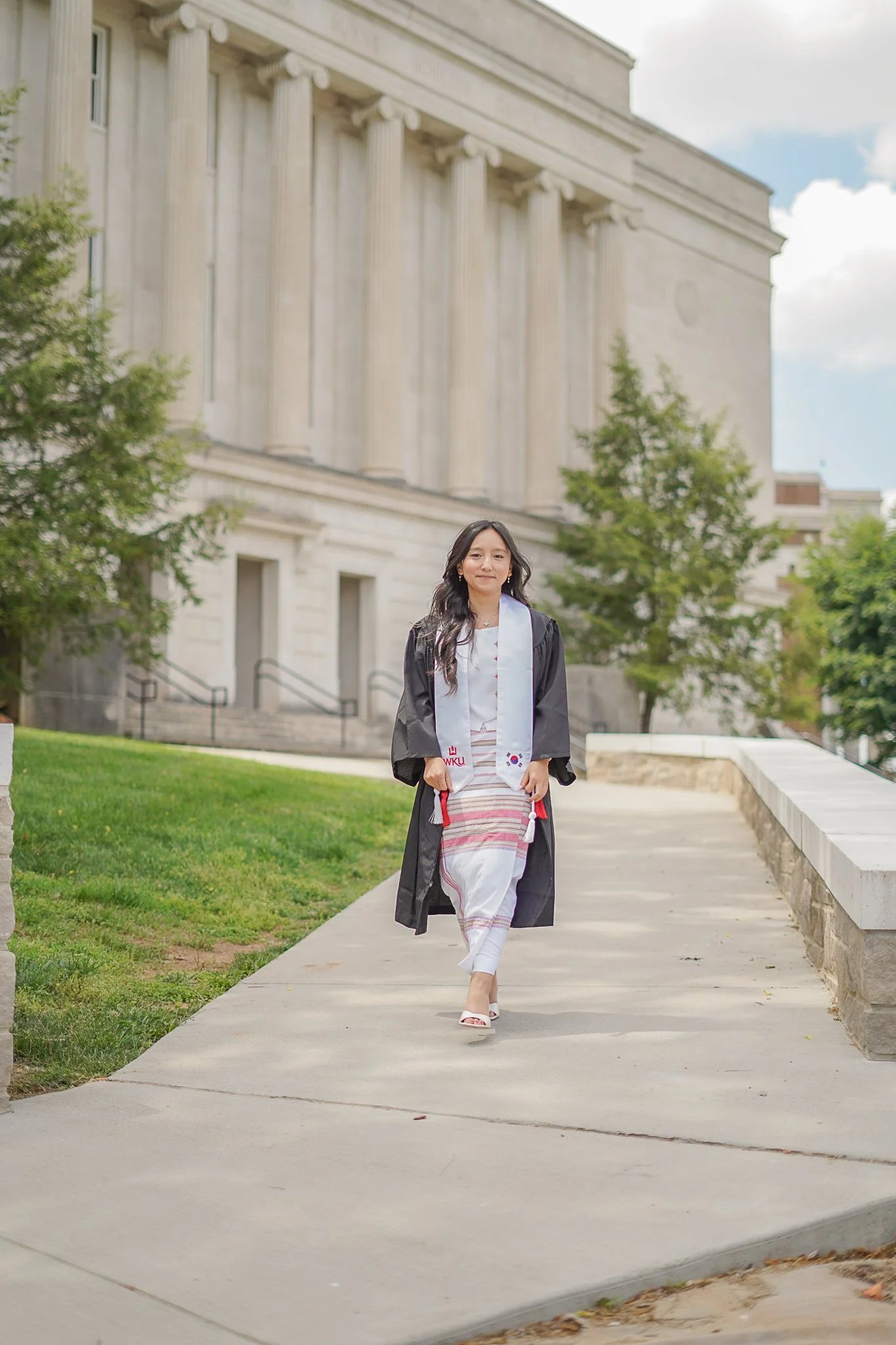 A woman in graduation attire walking outdoors in front of a large stone building with columns, trees, and a partly cloudy sky.