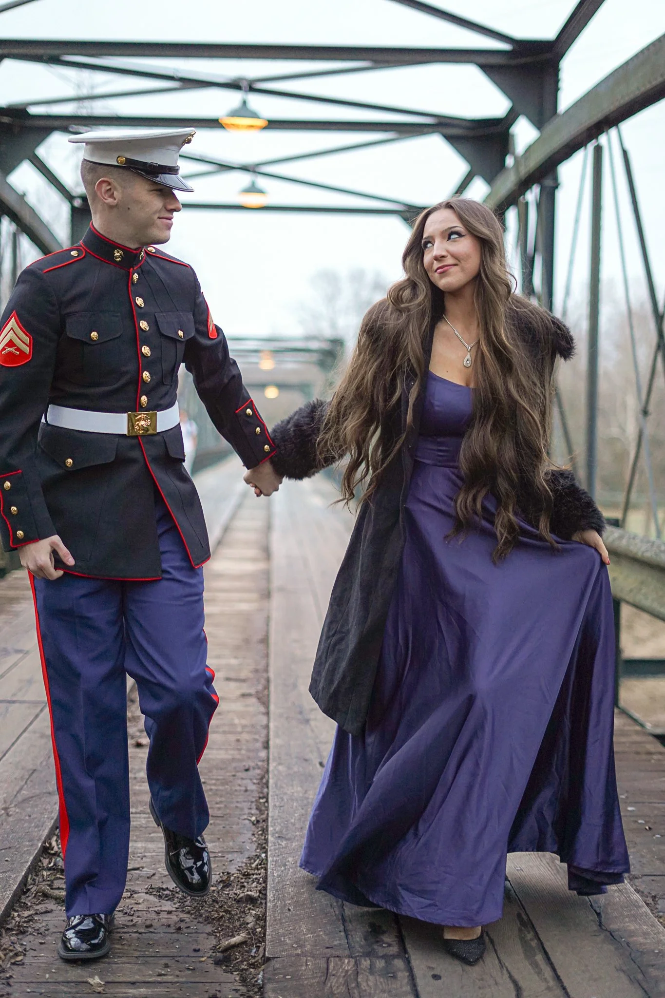 A young man in a United States Marine Corps dress uniform holding hands with a young woman in a purple dress and black fur coat on a bridge. The woman is looking at the man with a smile. The scene is outdoors on a cloudy day.