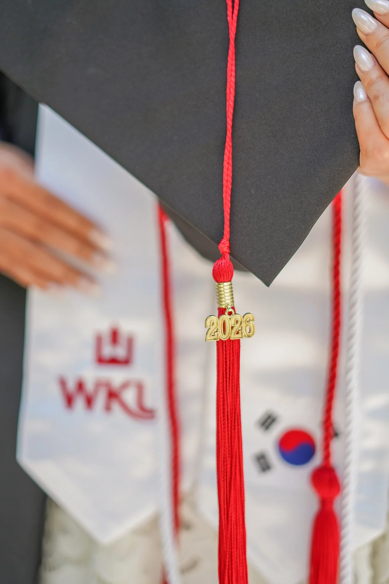 Close-up of a graduation cap with a red tassel featuring a small gold charm with the year '2026'; person with painted nails holding the cap.
