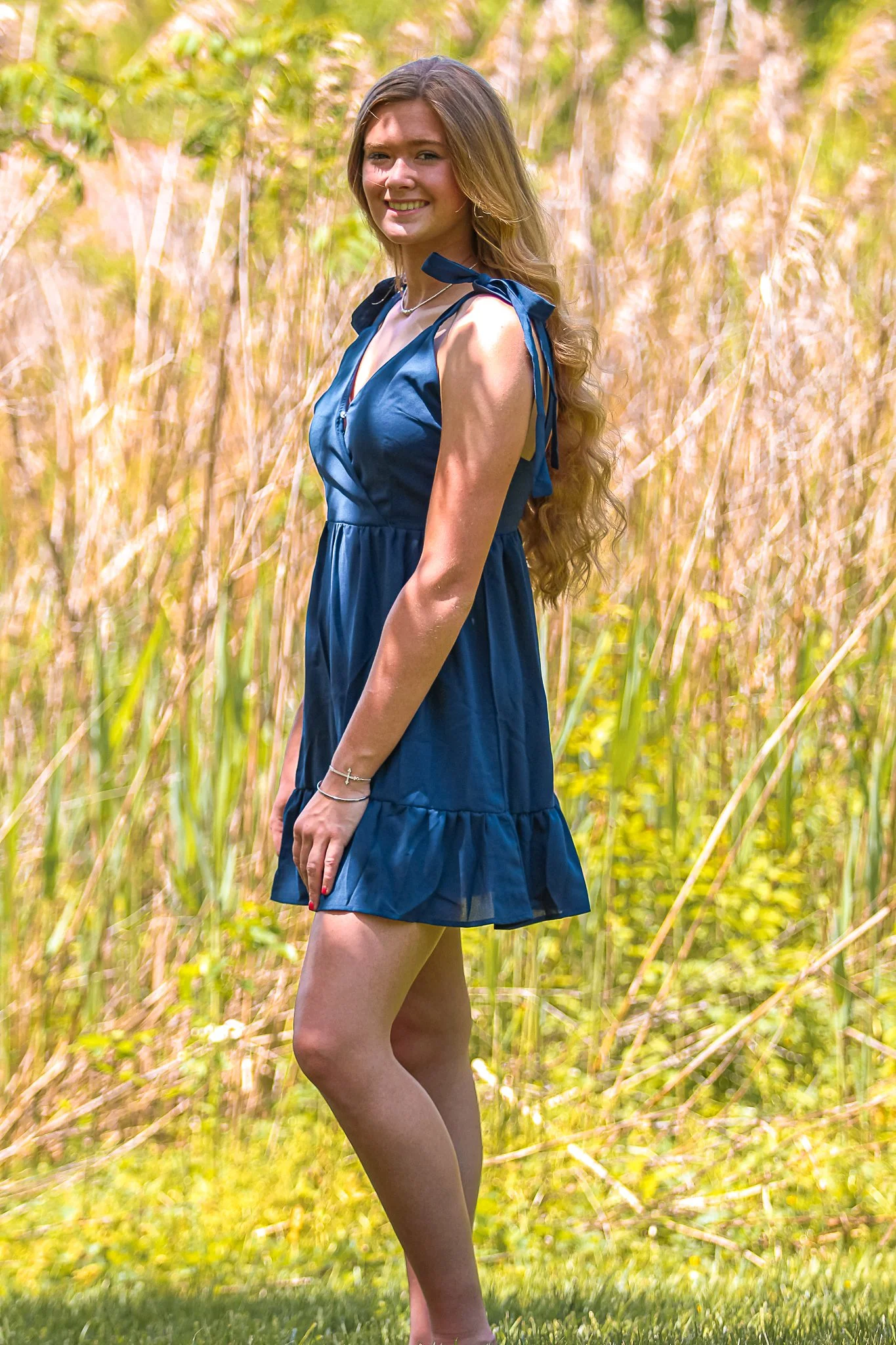 A young woman with long, wavy brown hair wearing a sleeveless blue dress with bow details stands outdoors in a field of tall grass and yellow-green foliage, smiling at the camera on a sunny day.