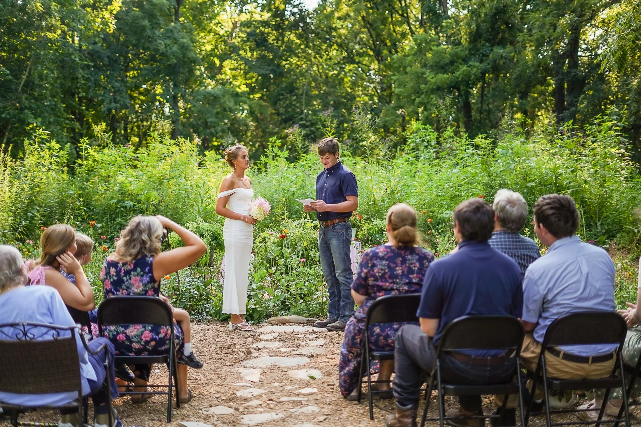 A wedding ceremony taking place outdoors in a lush green forest. The bride in a white dress holds a bouquet, standing next to the groom who reads from a paper. A group of people sits on chairs, watching the ceremony, surrounded by vibrant greenery an