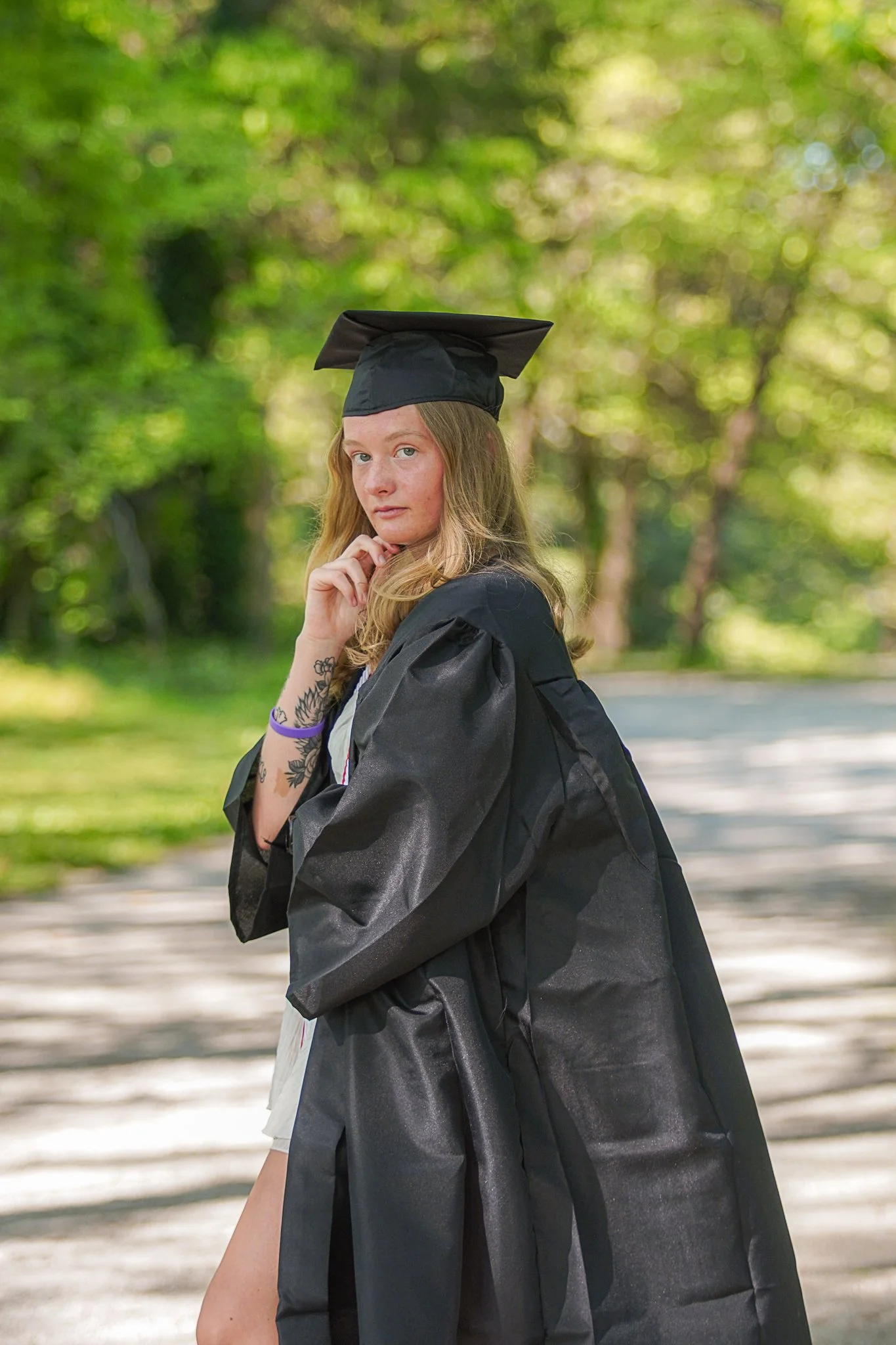 A young woman in a graduation cap and gown, standing outdoors on a sunny day with green trees in the background, looking at the camera with a thoughtful expression.