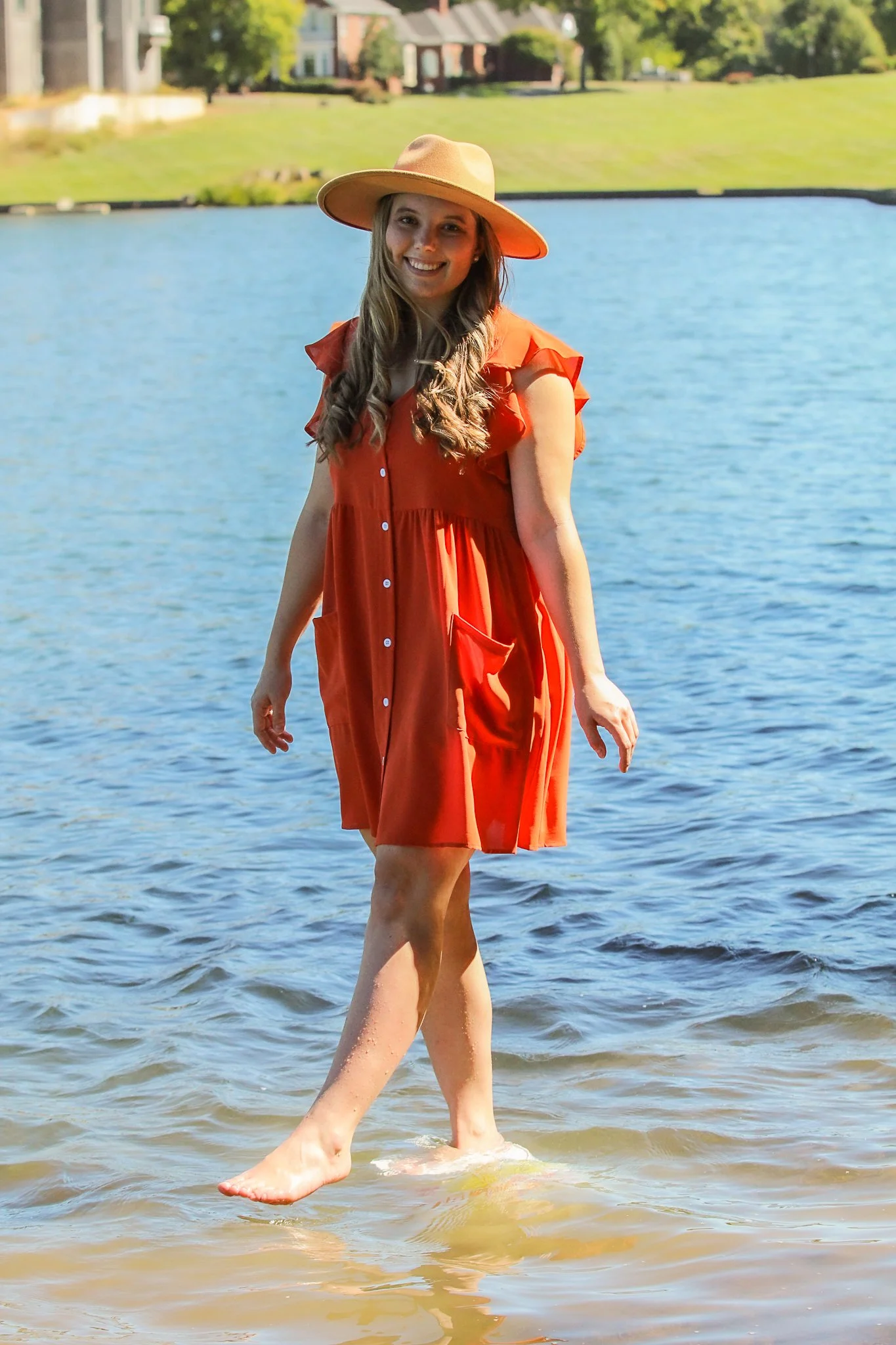 A woman in an orange dress and a wide-brimmed hat standing in the water by a lake, smiling at the camera.