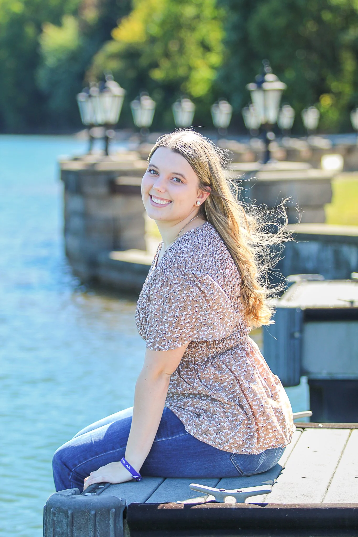 A young woman with long blond hair and a patterned dress sitting on a dock by the water, smiling.