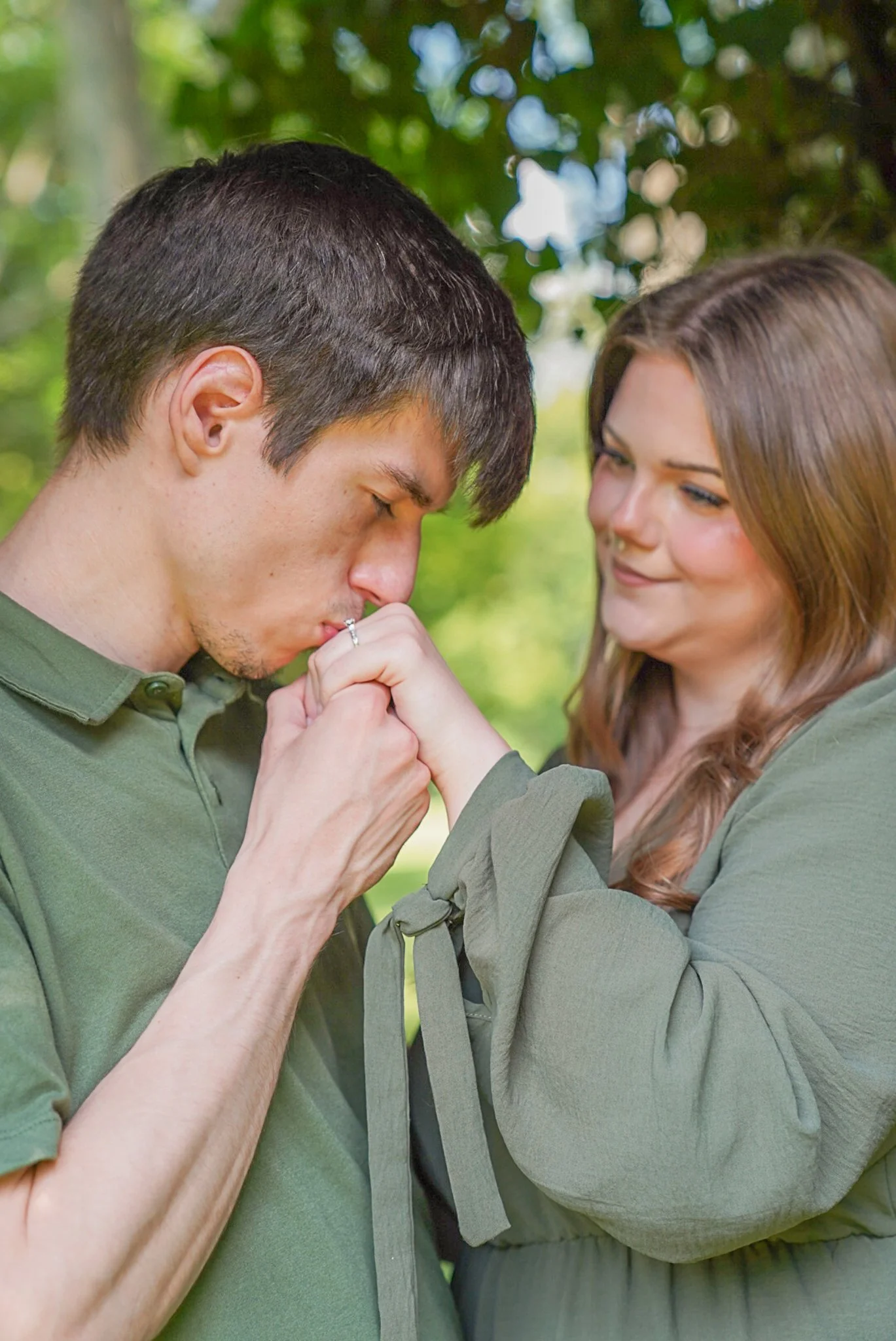 A man and a woman in an outdoor setting, with the man holding the woman's hand and kissing it, while both are smiling and looking at each other. They are wearing green shirts, and trees with green leaves are visible in the background.
