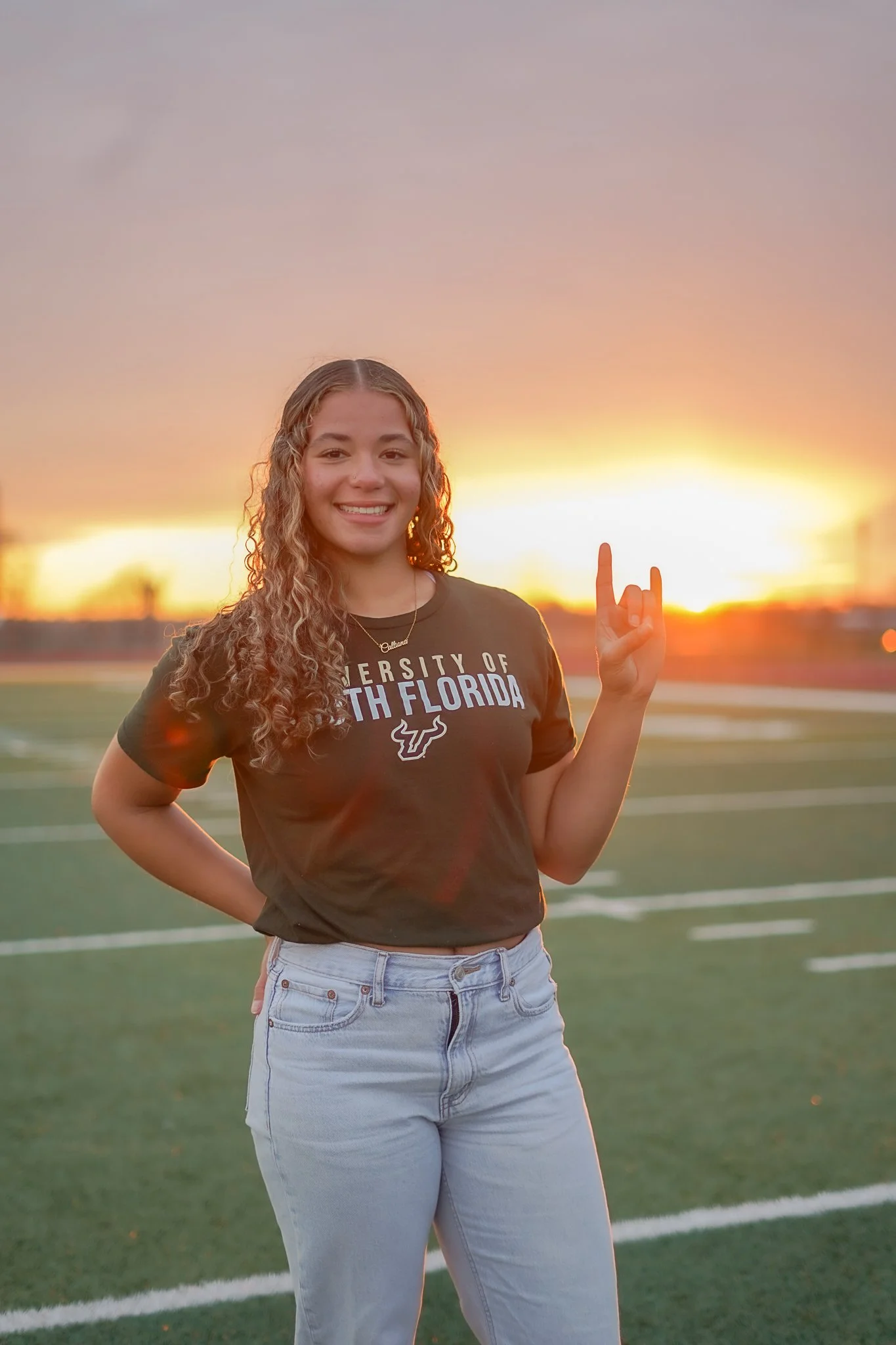 A woman standing on a football field during sunset, wearing a black T-shirt with 'University of South Florida' written on it, smiling and making a hand gesture.