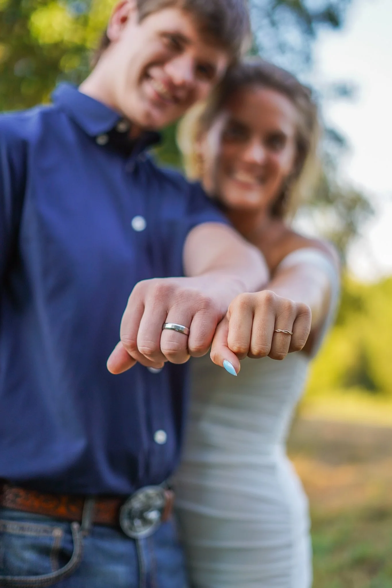 Close-up of a happy couple showing their wedding rings on their outstretched hands, with greenery in the background.