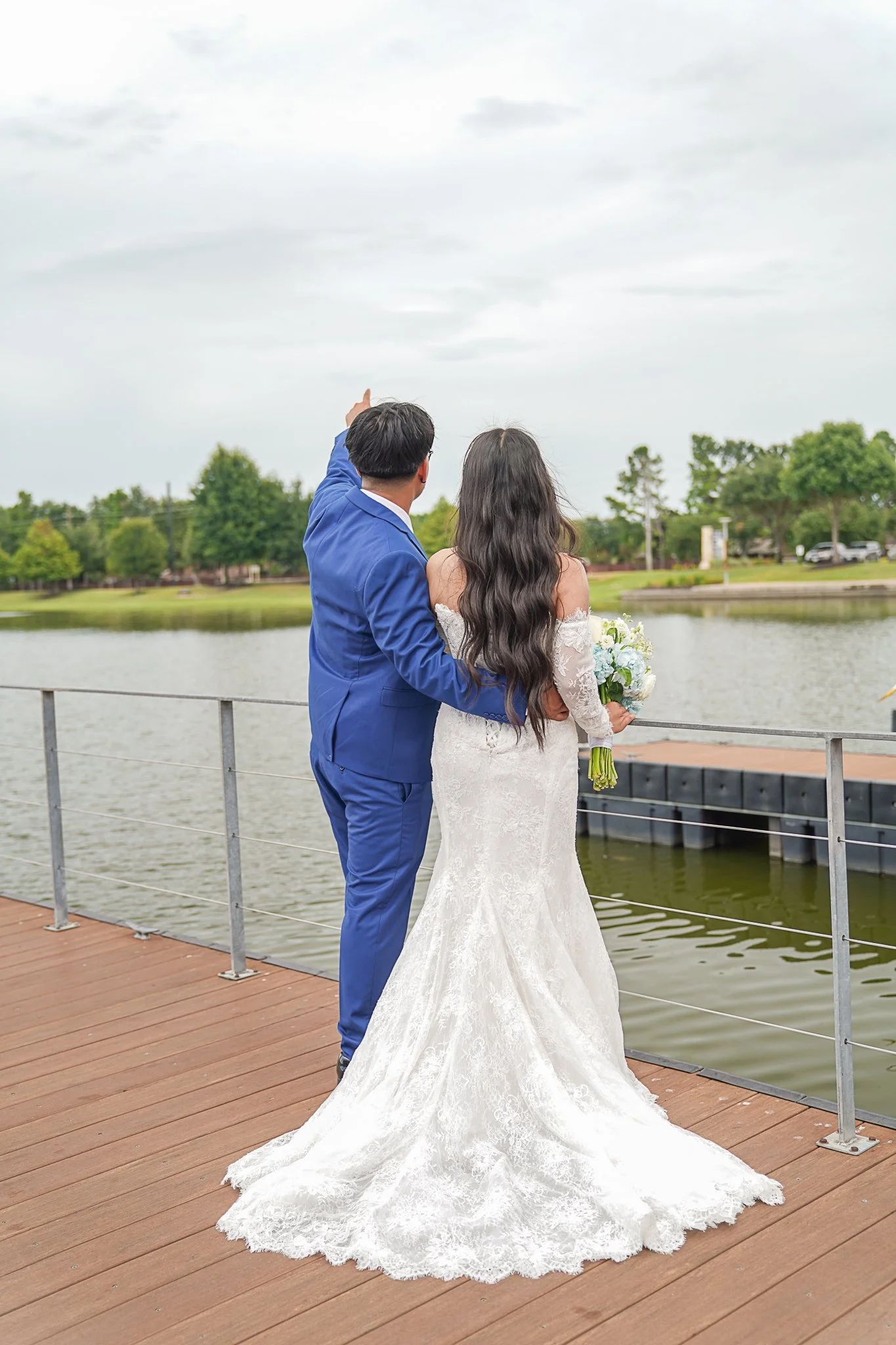 A bride and groom stand on a wooden dock by a lake, with the groom wearing a blue suit and the bride in a white wedding gown holding a bouquet, as they look at the cloudy sky.