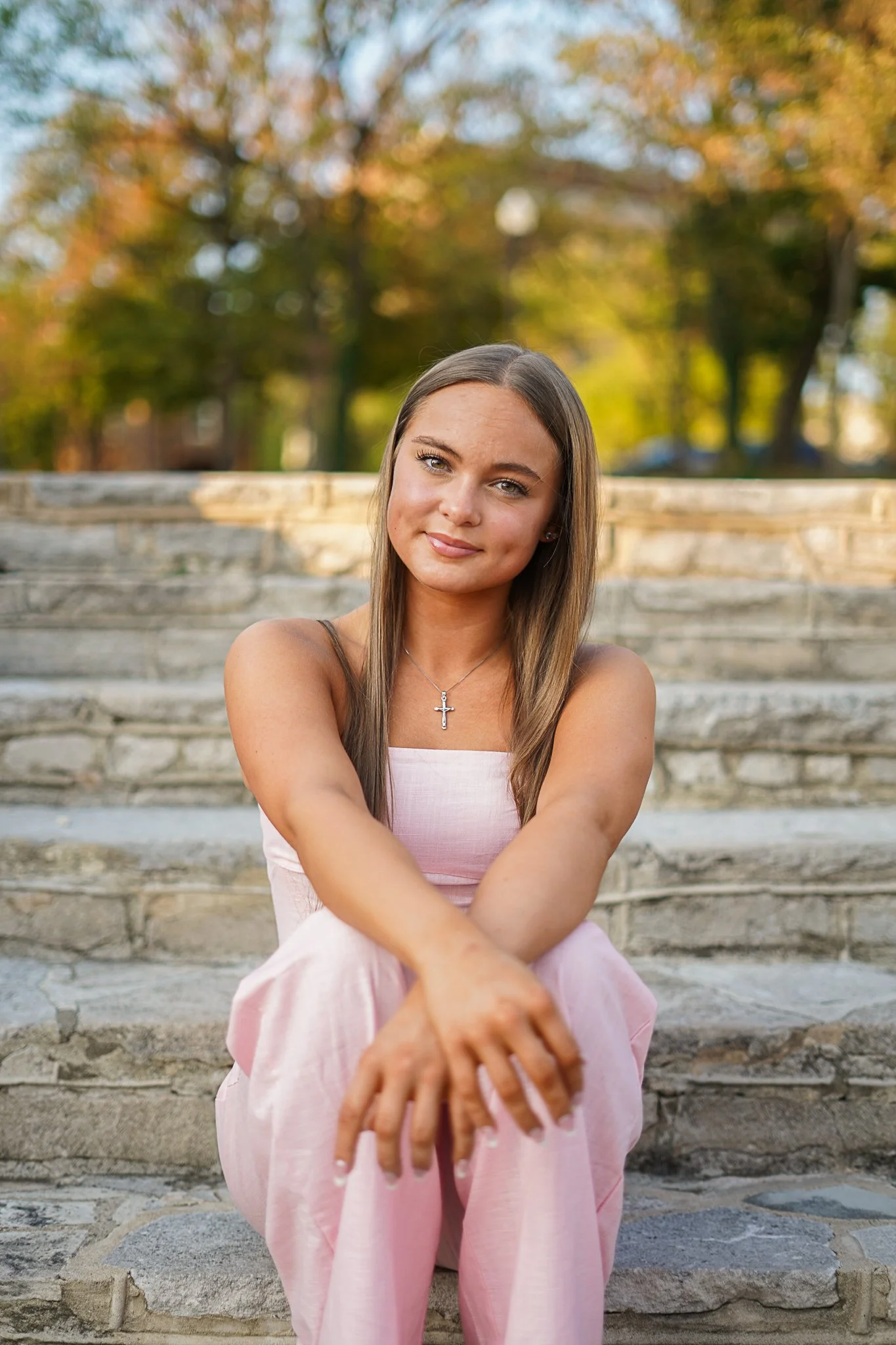 A young woman with long brown hair sitting on stone steps outside in a park during autumn, wearing a pink dress and a cross necklace, smiling at the camera.