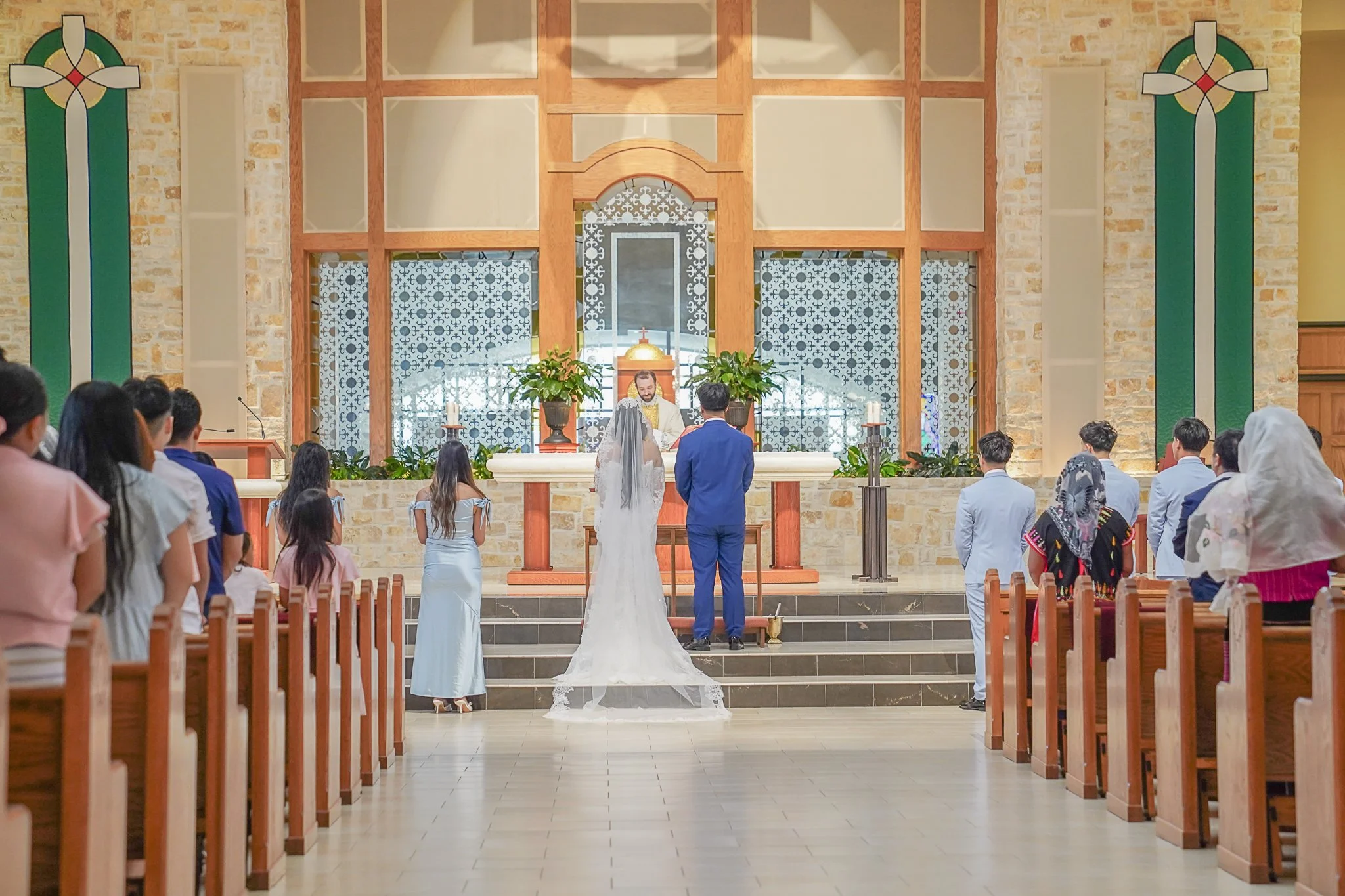 A wedding ceremony inside a church with a bride and groom standing at the altar facing a priest. Guests are seated in pews on either side, observing the ceremony. The church has stone walls, stained glass windows, and decorative plants near the altar