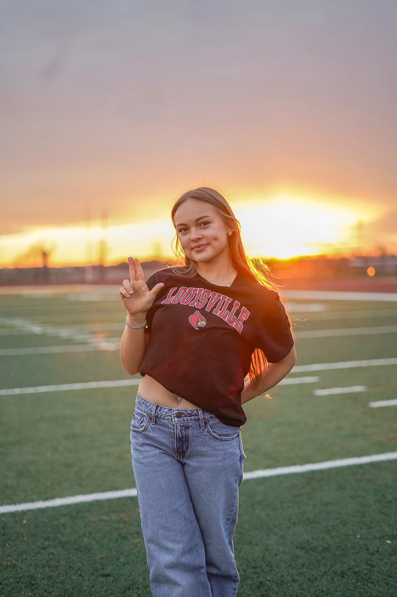 A young woman standing outdoors on a sports field during sunset, wearing a Louisville T-shirt and high-waisted jeans, making a peace sign with her right hand and smiling.
