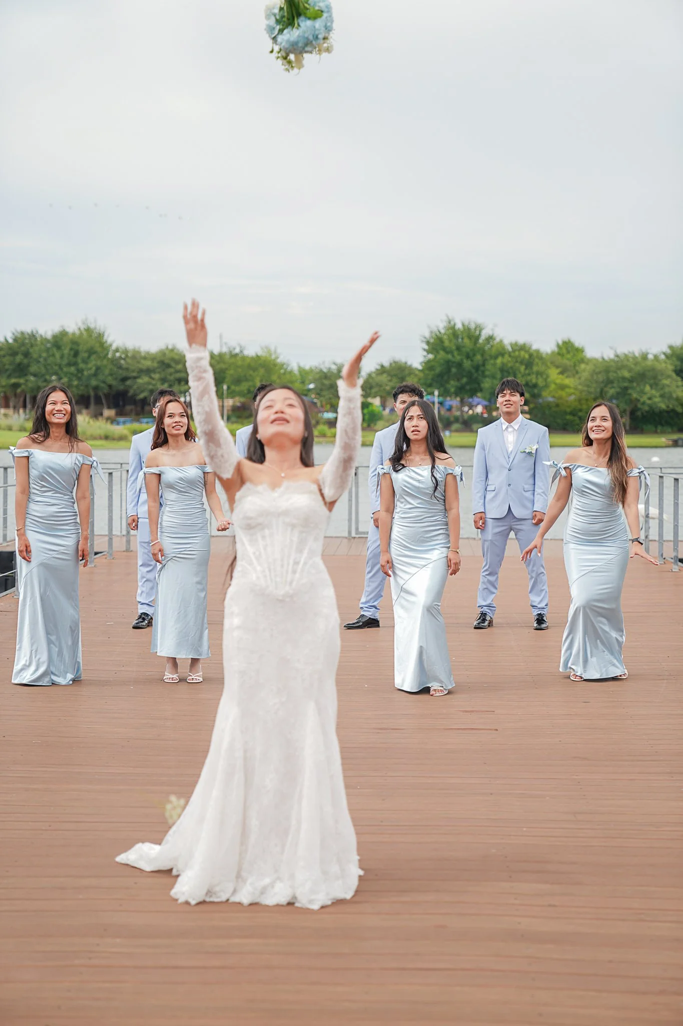 Bride in a white wedding dress releasing a bouquet into the air as bridesmaids and groomsmen watch outdoors by a lake.